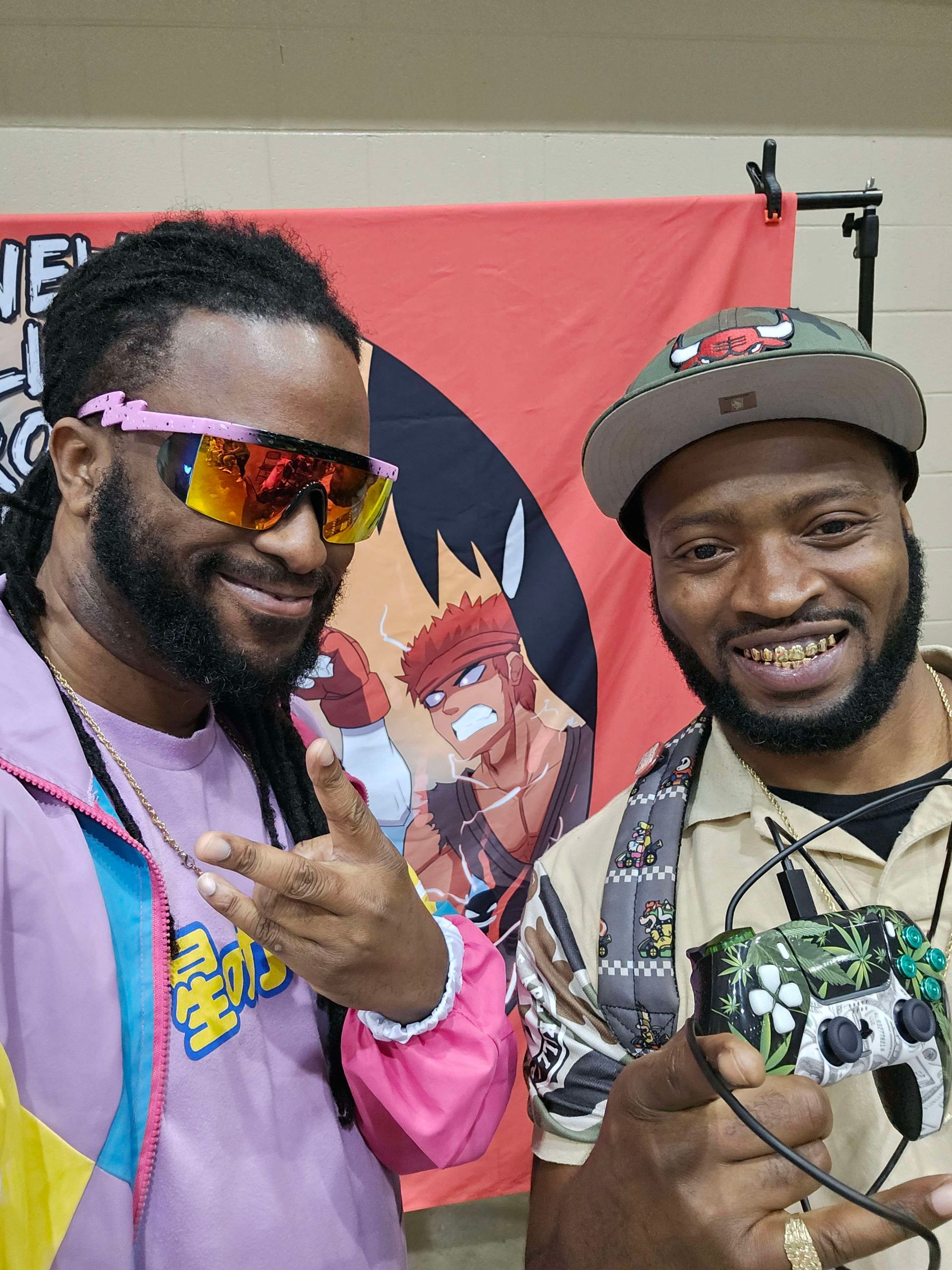 Two men smiling, one in pink jacket, sunglasses; other with controller, Chicago Bulls hat. Backdrop with cartoon.