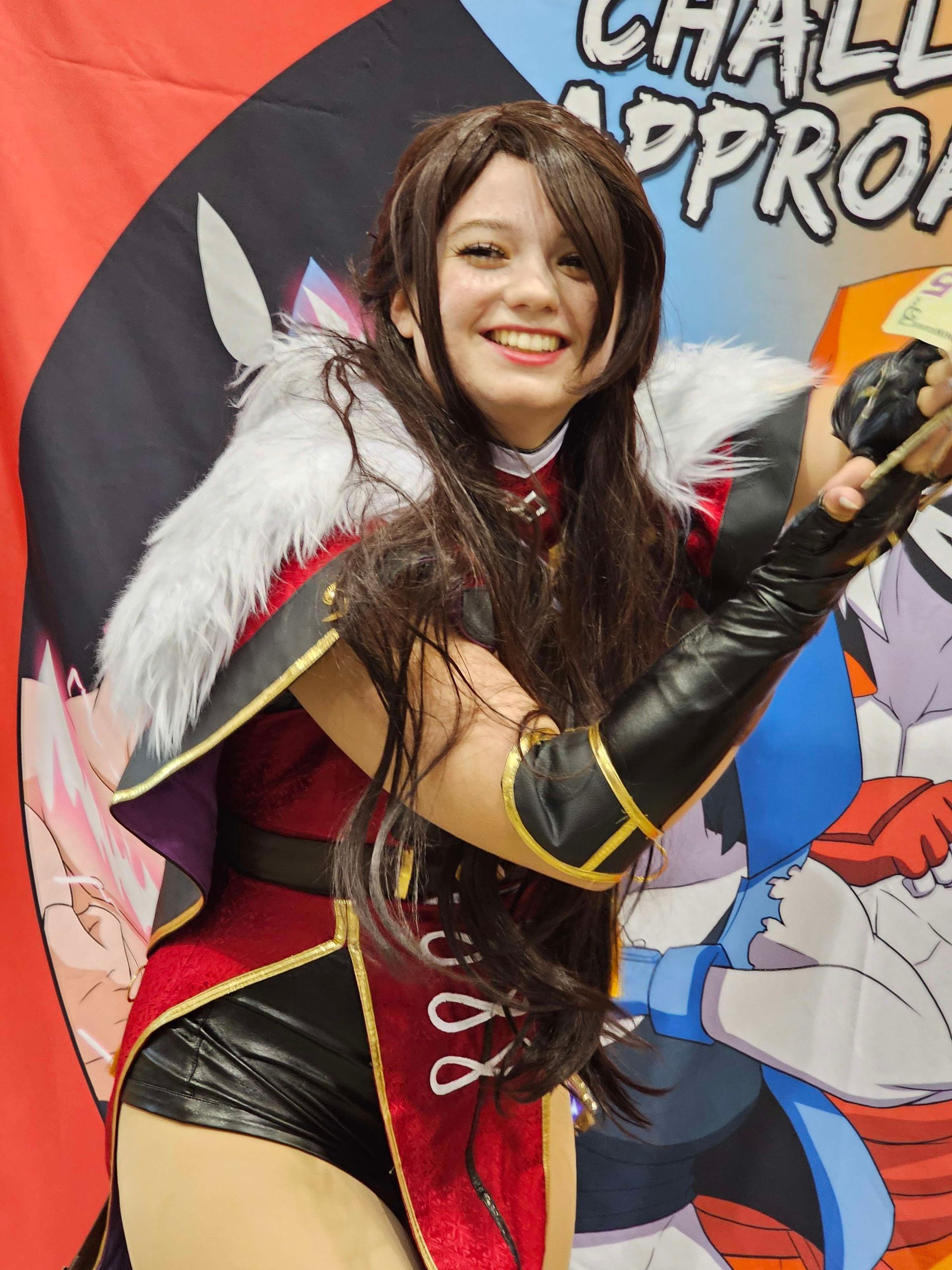 Cosplayer in red and black outfit smiles, arms raised, against a character banner.