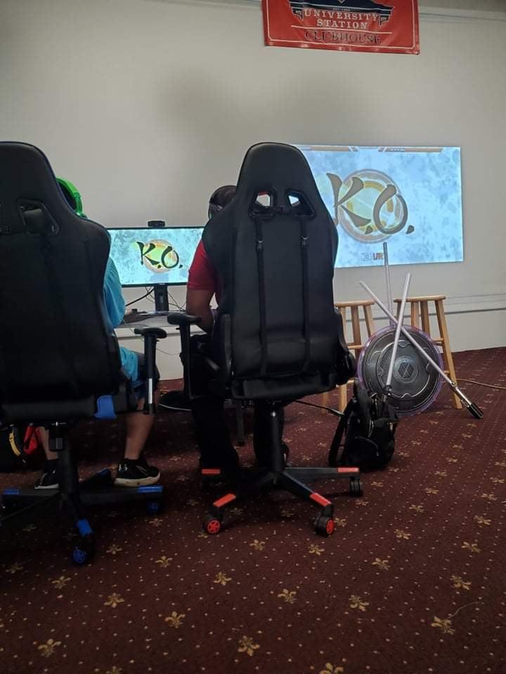 People seated at gaming computers, screens displaying logos. Chairs and equipment are in a room, with a fan and poster.