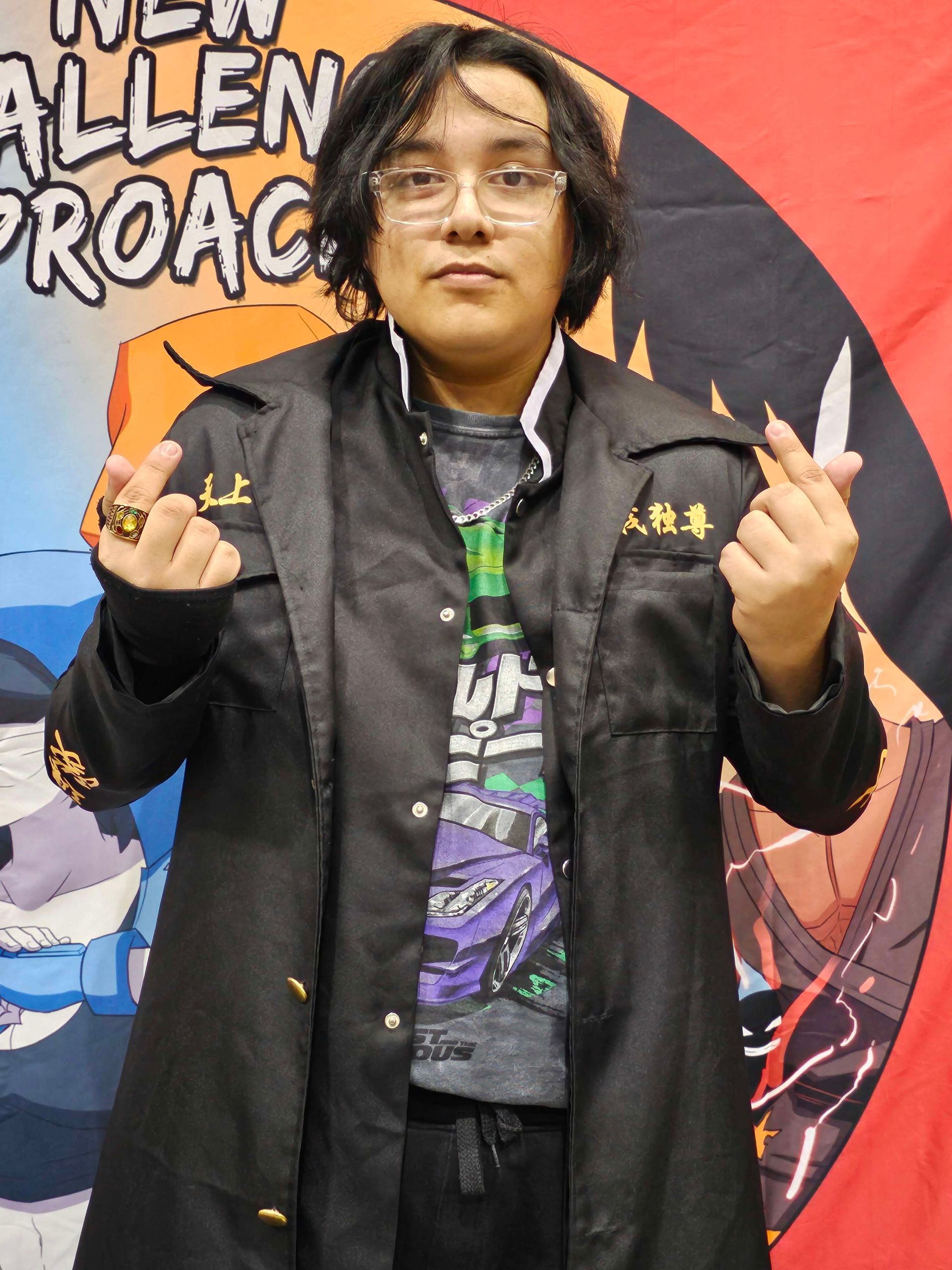 Person in black jacket makes a heart shape with fingers, standing in front of a banner.