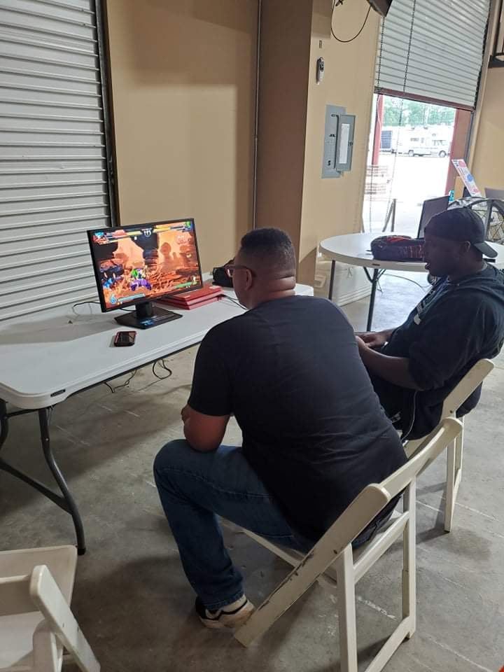 Two people seated, watching a video game on a monitor at a table in a room with a garage-style door.