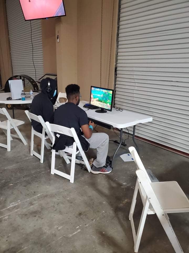 Two people playing video games at a table. A monitor displays gameplay. White chairs and a garage door are visible.