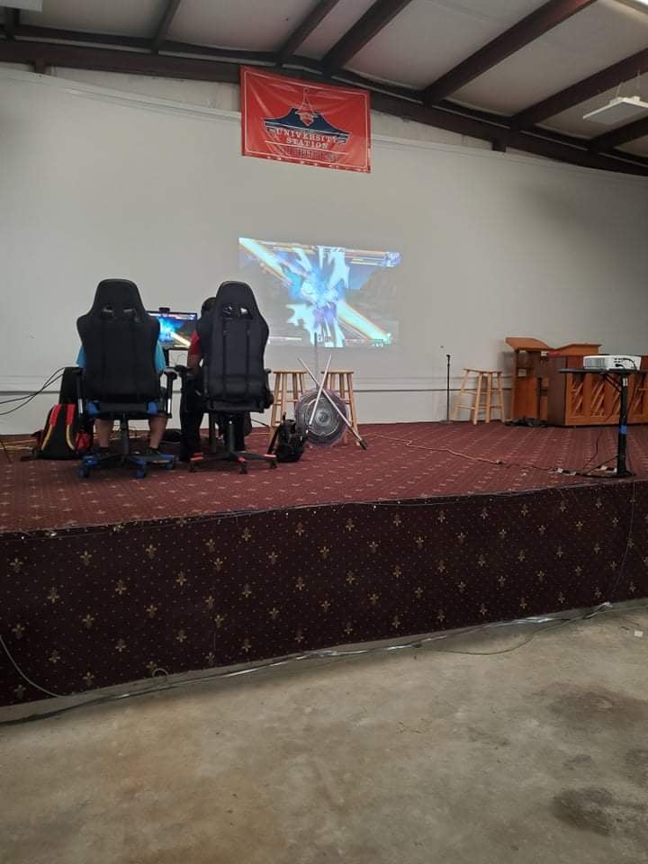 Two people gaming on a stage. A screen displays gameplay; a red banner is above.