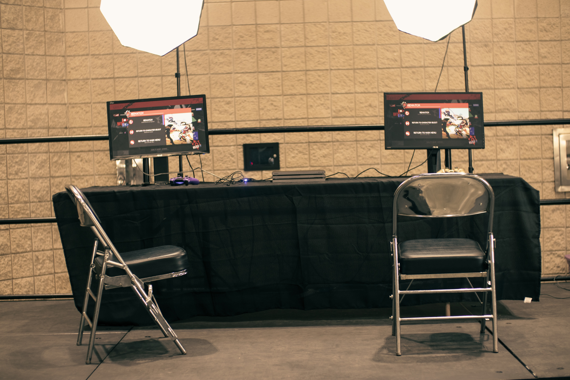 Two monitors on a table with a black cloth. Two chairs in front. Two overhead lights.