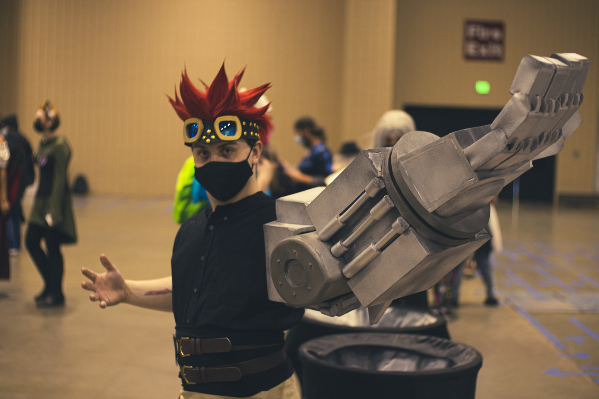 Cosplayer with red spiky hair, wearing goggles and a large robotic arm, posing at a convention.