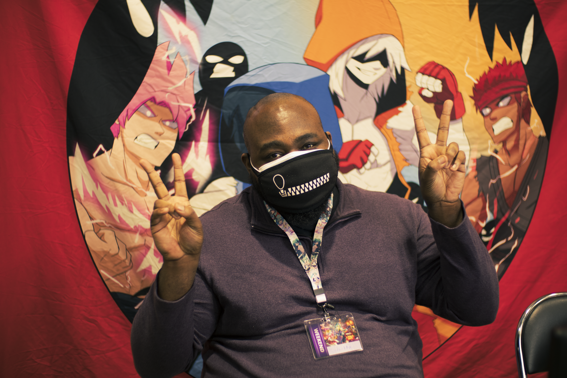 Person wearing mask with peace signs, seated in front of anime-style backdrop.