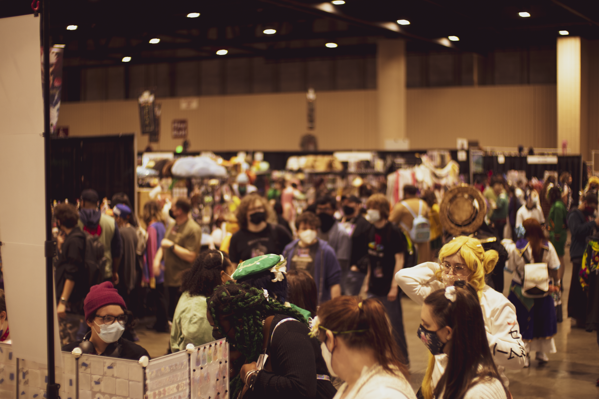 Crowd at a convention, people browsing vendor booths. Many wear masks, some cosplay. Inside a large hall with booths.