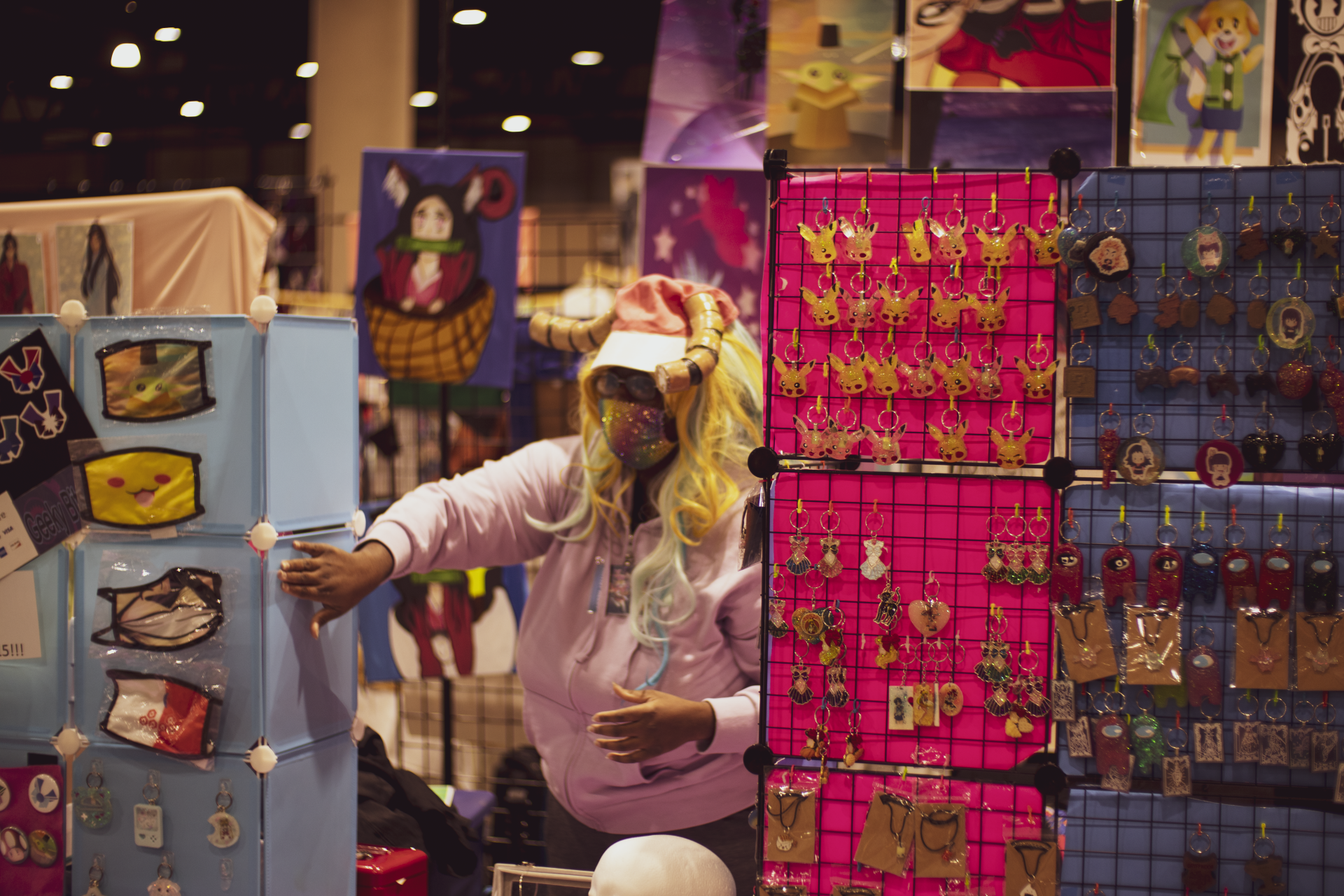 Person arranging jewelry at a vendor booth in a convention hall, with colorful art on display.