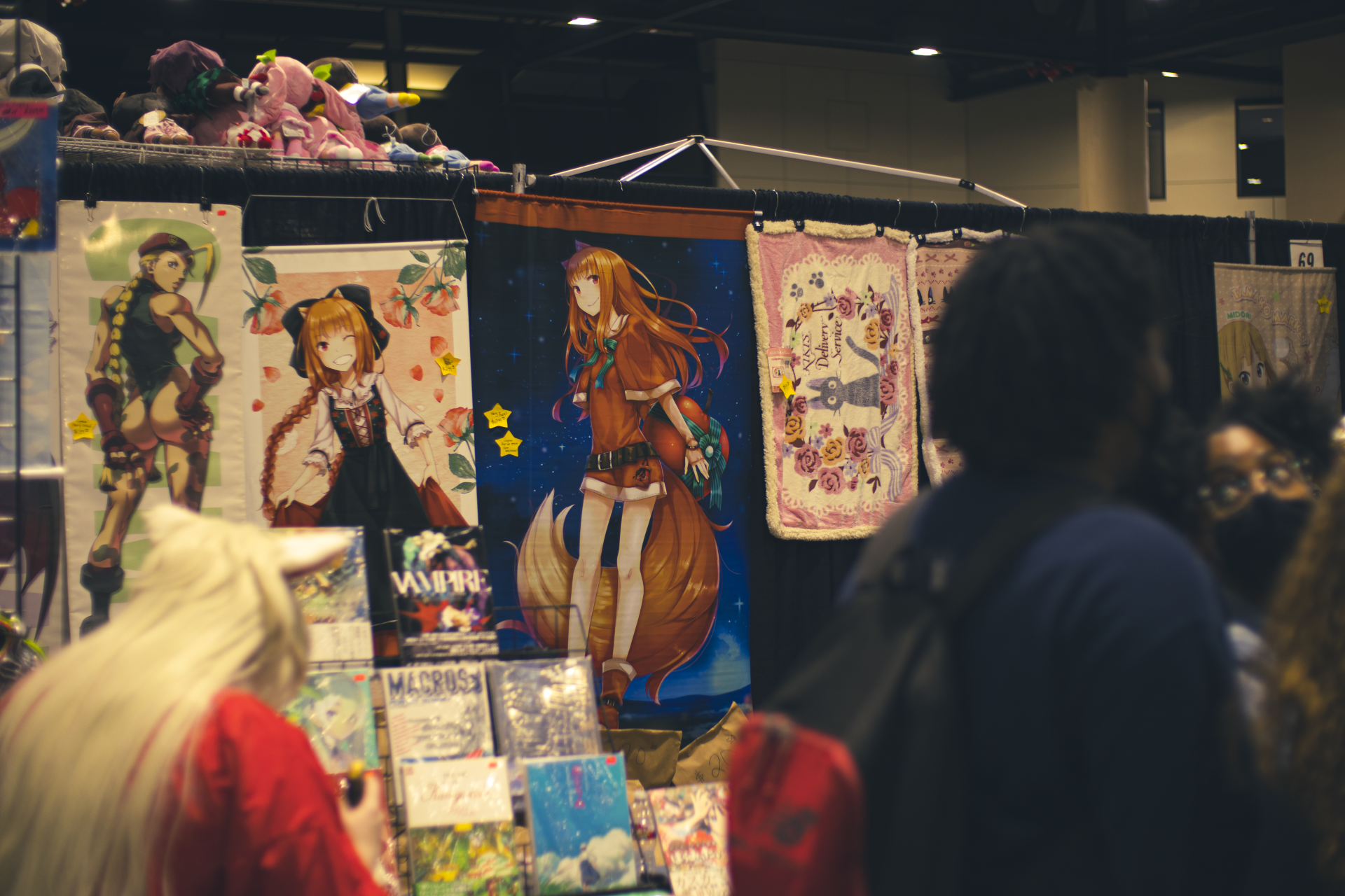 Anime art booth at a convention. Posters of anime characters and other goods displayed. Several people browse the booth.