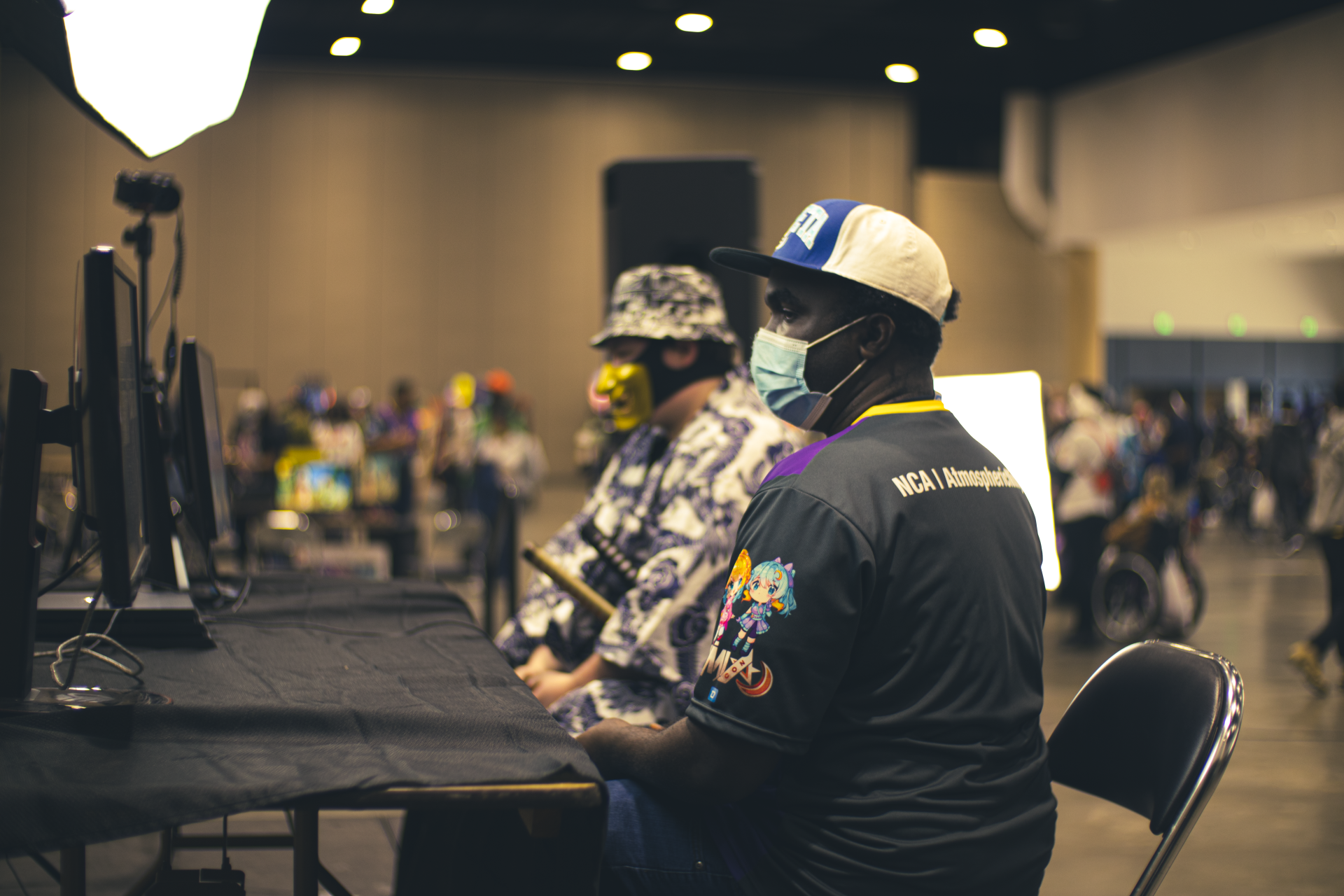 Two people wearing masks and hats at computer monitors. Indoor event with a crowd.