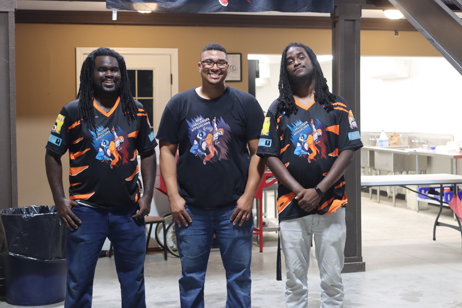 Three men standing inside, two wearing matching black jerseys with orange accents, one in a graphic black t-shirt.