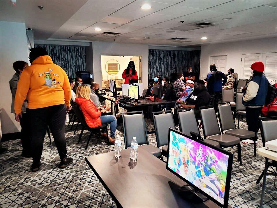 People in a room with computers, a monitor showing anime. Some are seated, some standing, some wearing colorful clothing.