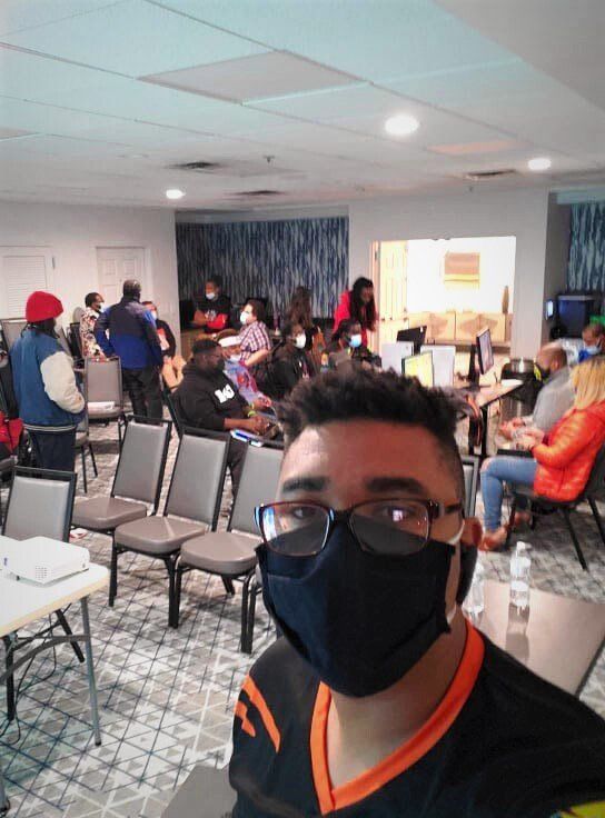 A person in a mask takes a selfie in a room with people sitting at tables and chairs; a projector on a table.