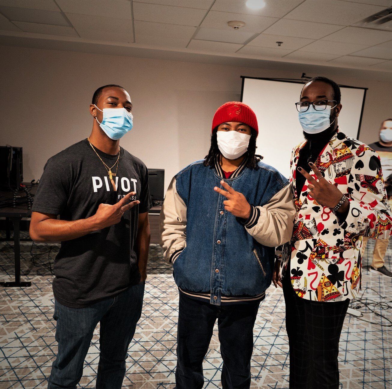 Three people wearing masks pose indoors, making the peace sign.