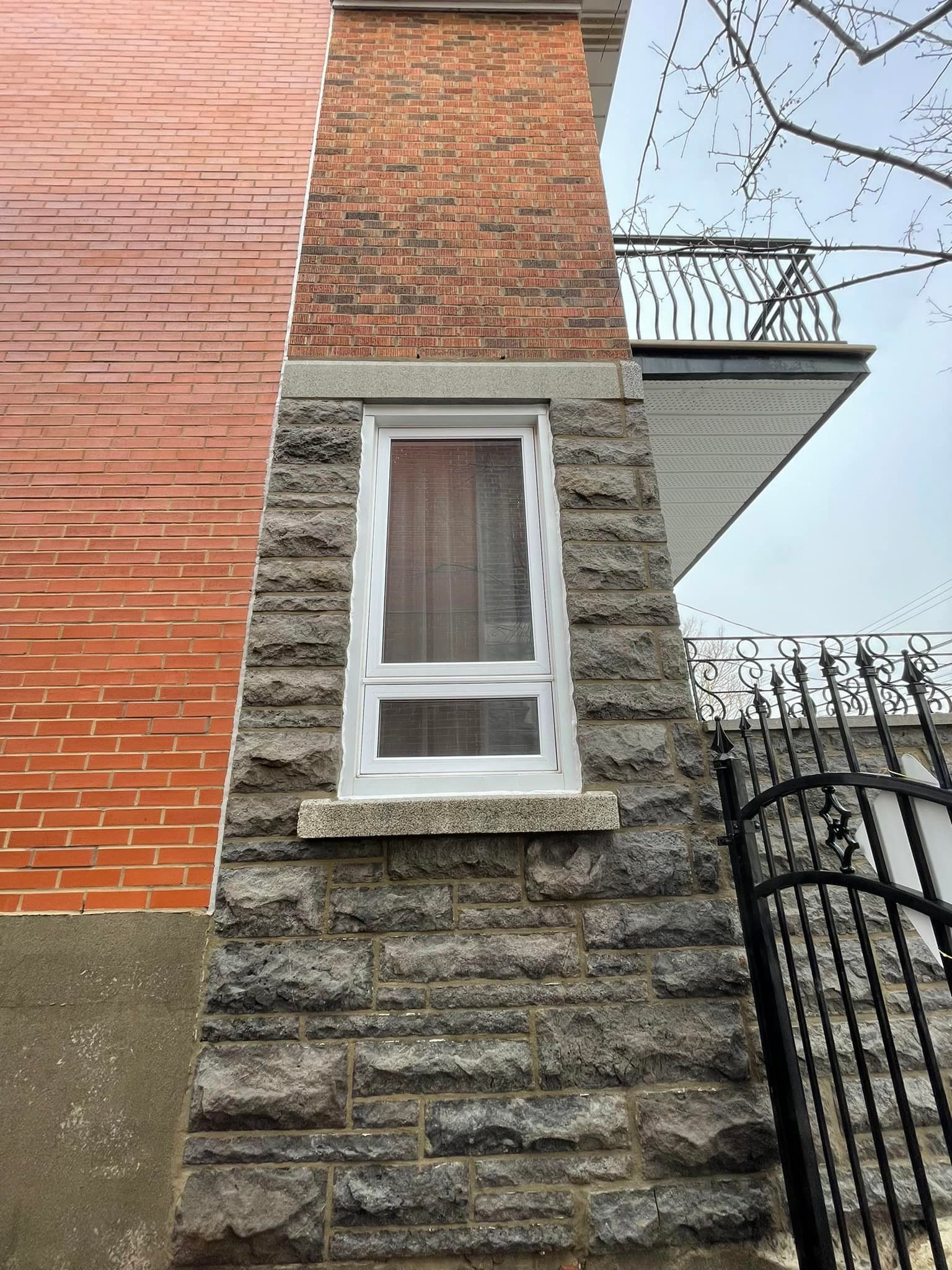 Bâtiment extérieur avec un pilier et une fenêtre recouverts de pierre. Mur de briques à gauche et rambarde métallique.