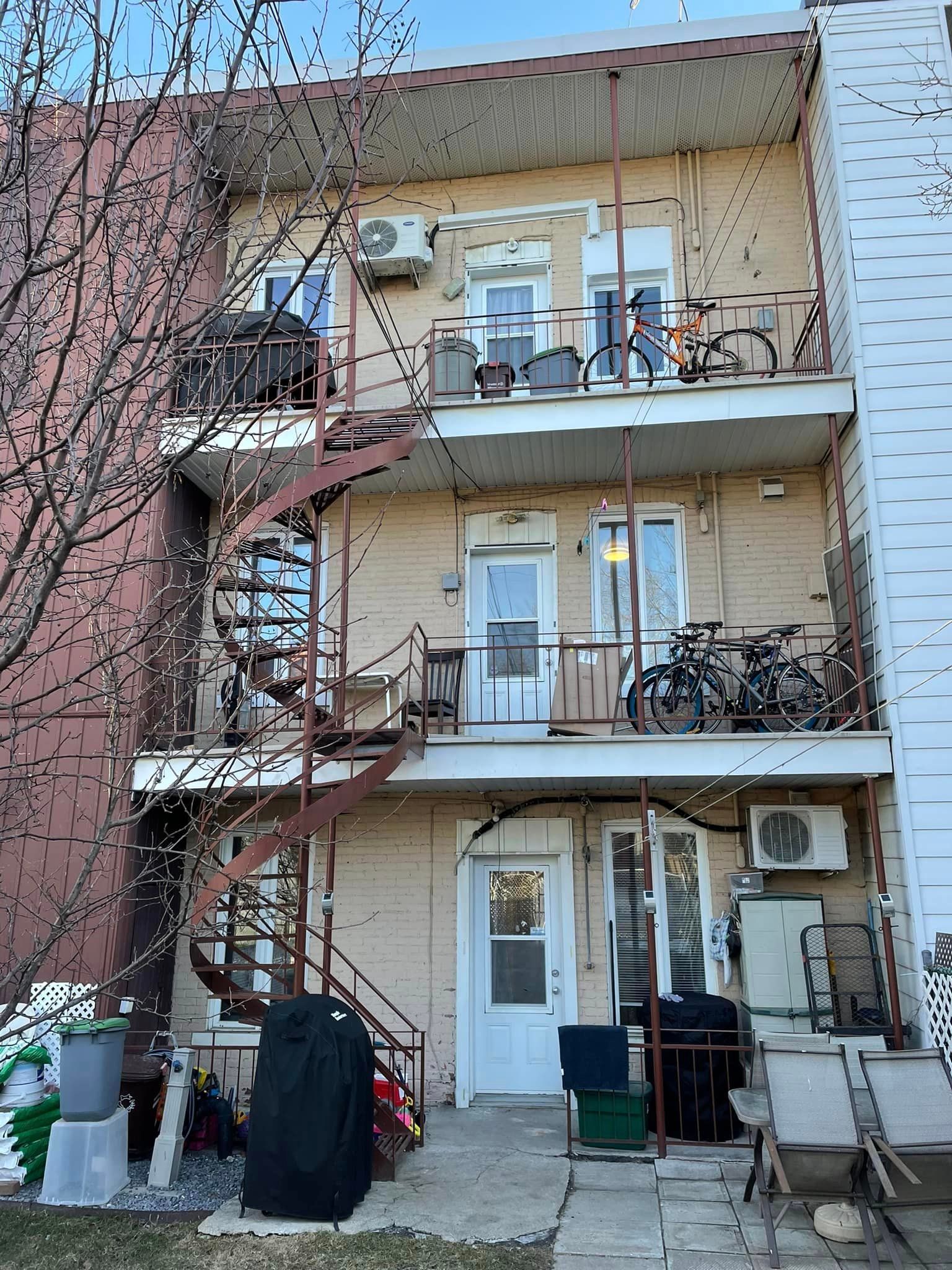 Arrière d'un immeuble de trois étages avec escaliers de secours et balcons. Murs beiges, climatiseurs et divers objets sur les balcons.