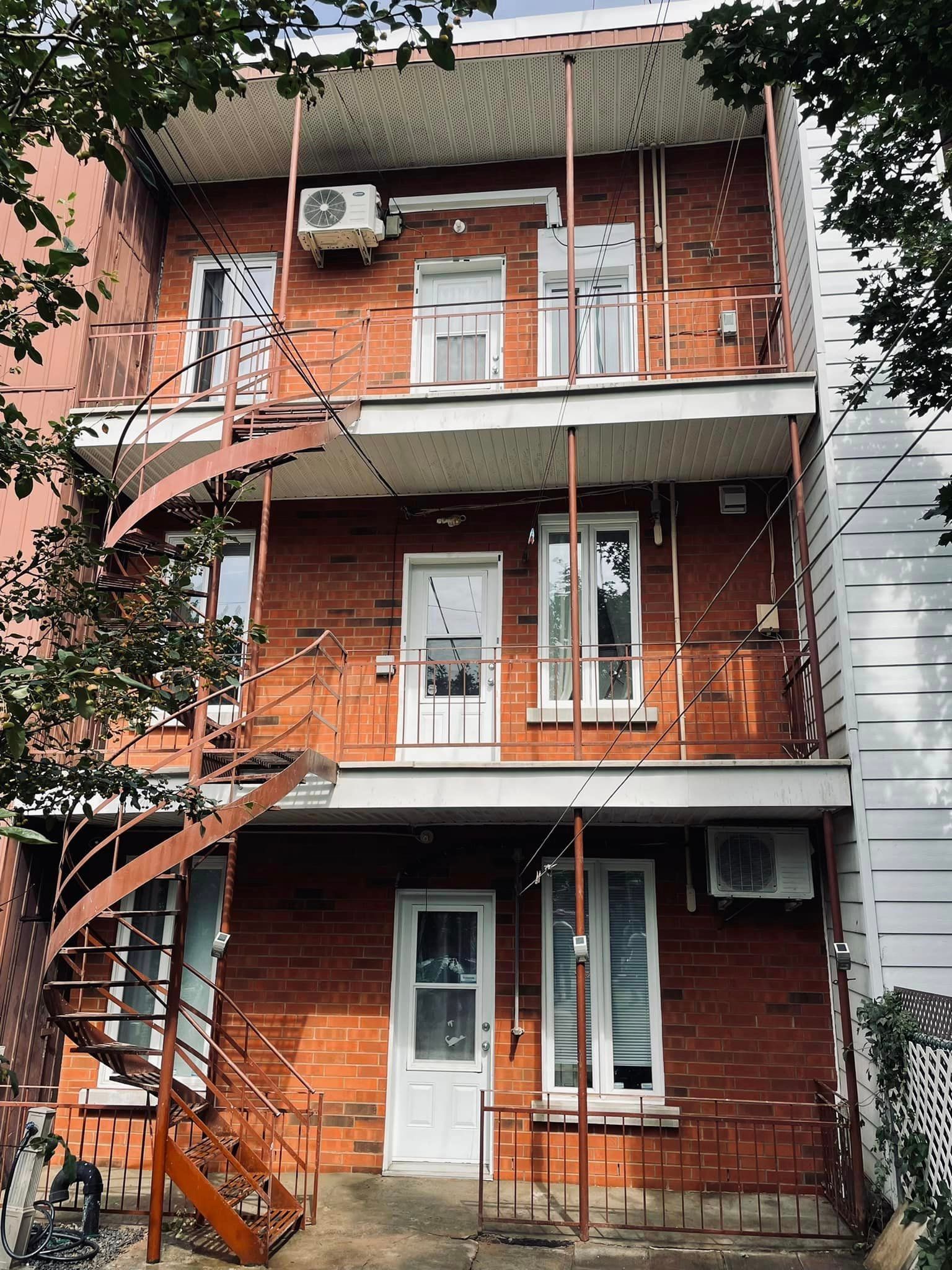 Bâtiment en briques rouges avec escalier extérieur en colimaçon, balcons et climatiseurs.
