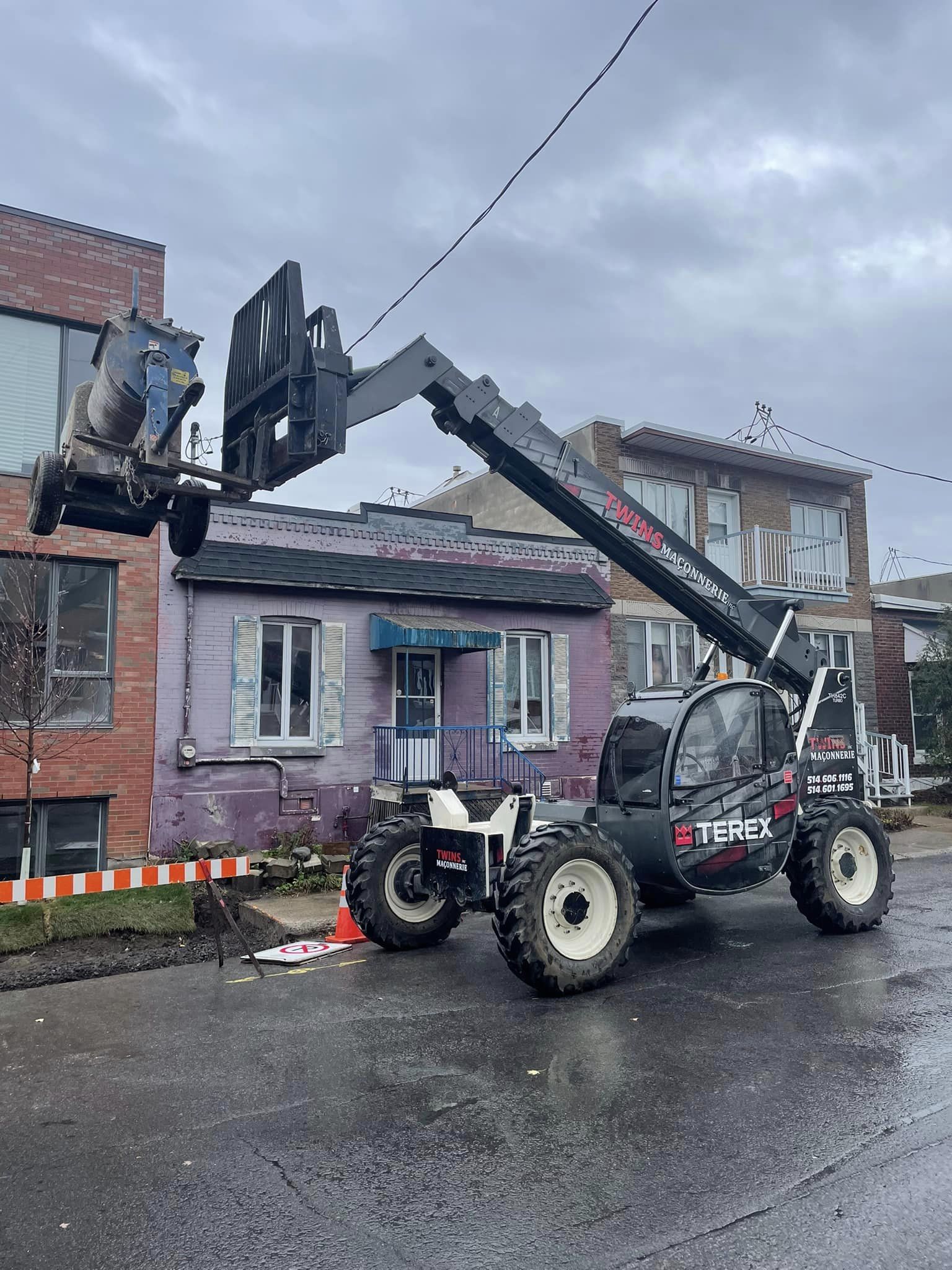Un chariot télescopique démolit la façade d'un bâtiment ; une structure en briques et une maison violette se trouvent en arrière-plan ; ciel couvert.