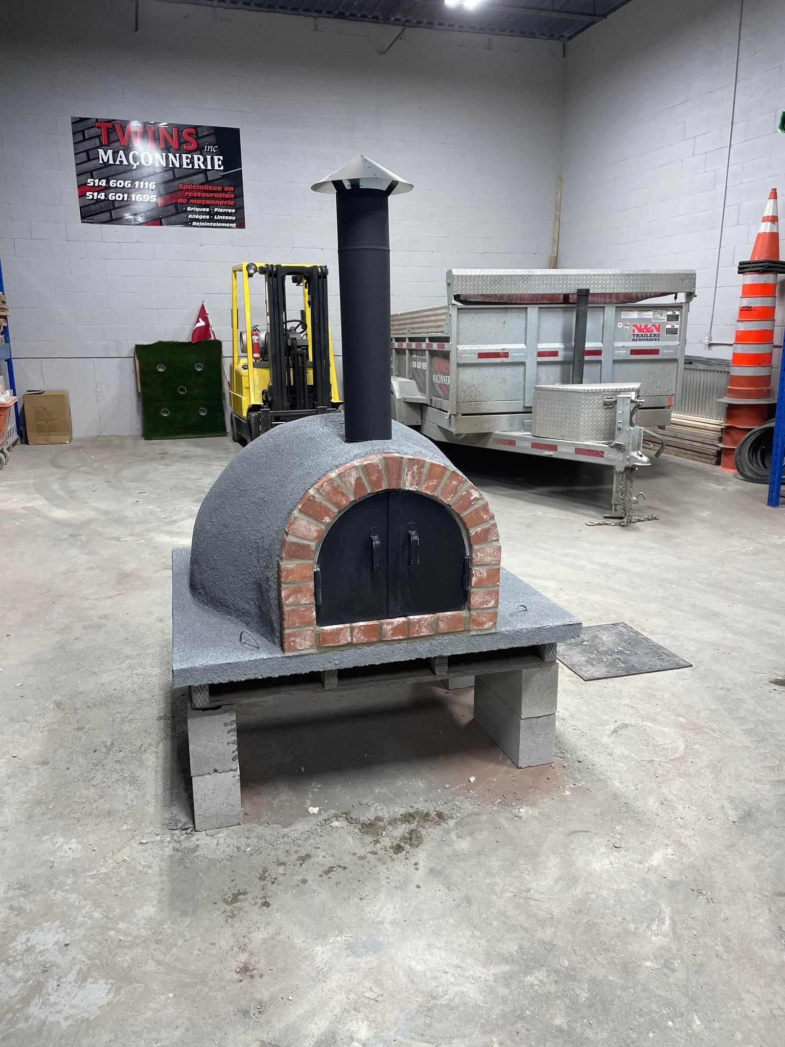 Four à pizza en briques sur une plateforme en béton gris soutenue par des parpaings dans un entrepôt.
