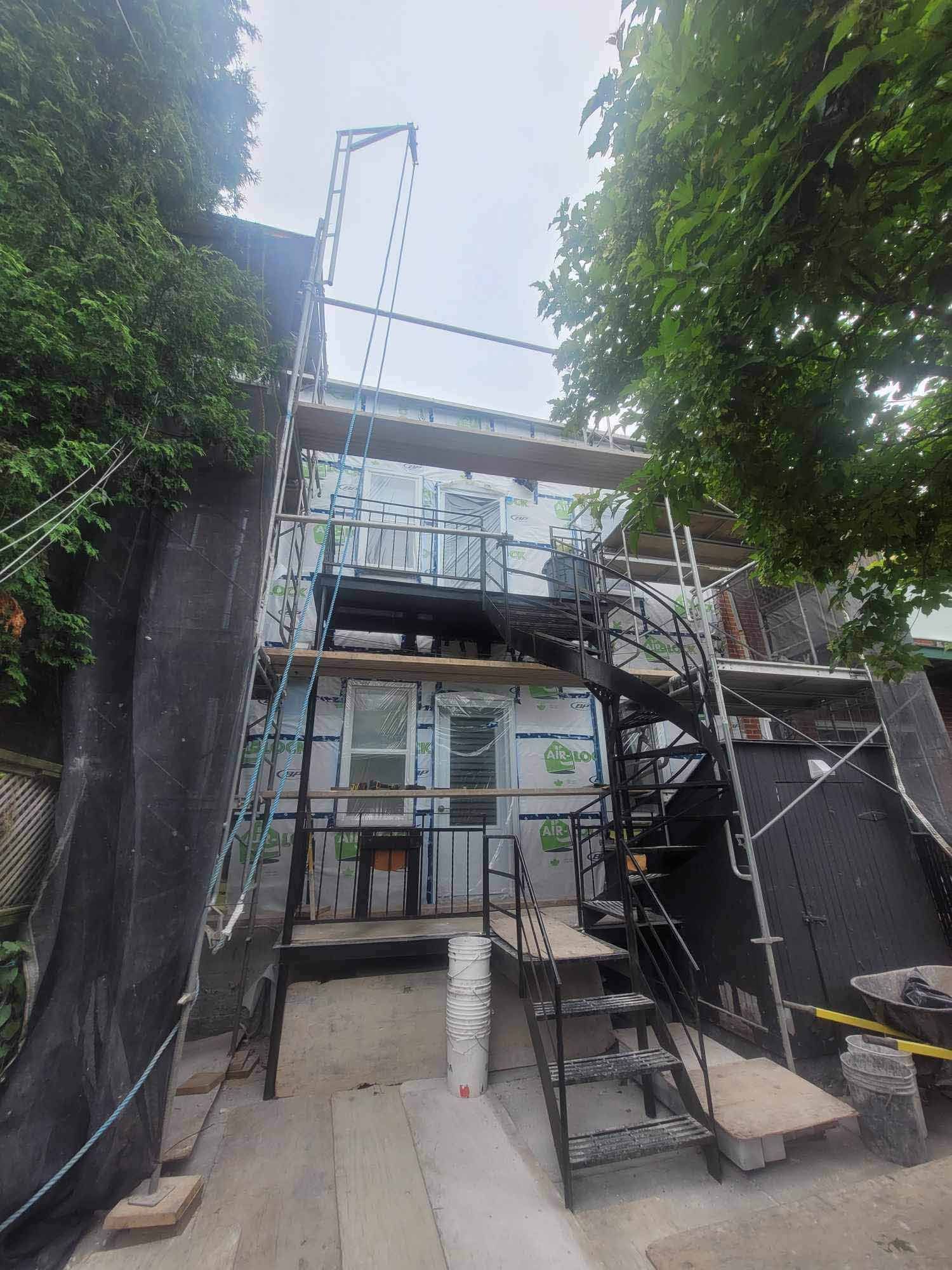 Extérieur d'un bâtiment en rénovation avec échafaudages et escalier métallique, entouré d'arbres.
