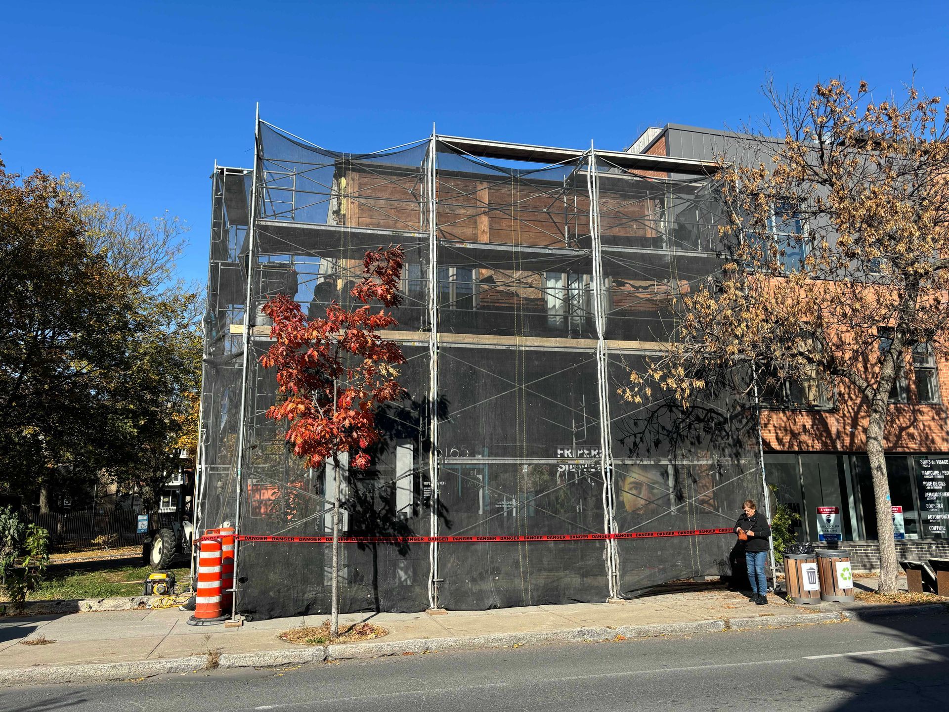 Bâtiment en construction, recouvert d'échafaudages et de filets noirs. Un arbre rouge et un ciel dégagé sont visibles.