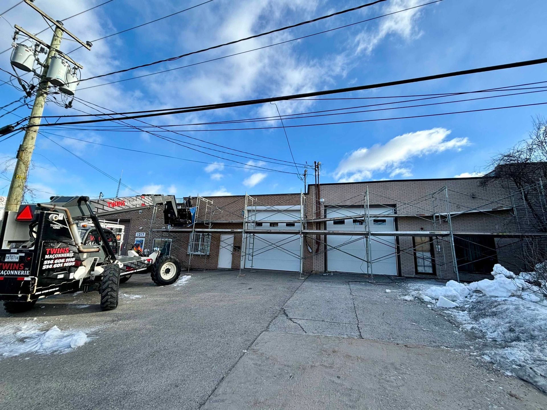 Vue d'un bâtiment avec des échafaudages, deux portes de garage et un tracteur dans une ruelle enneigée sous un ciel bleu.