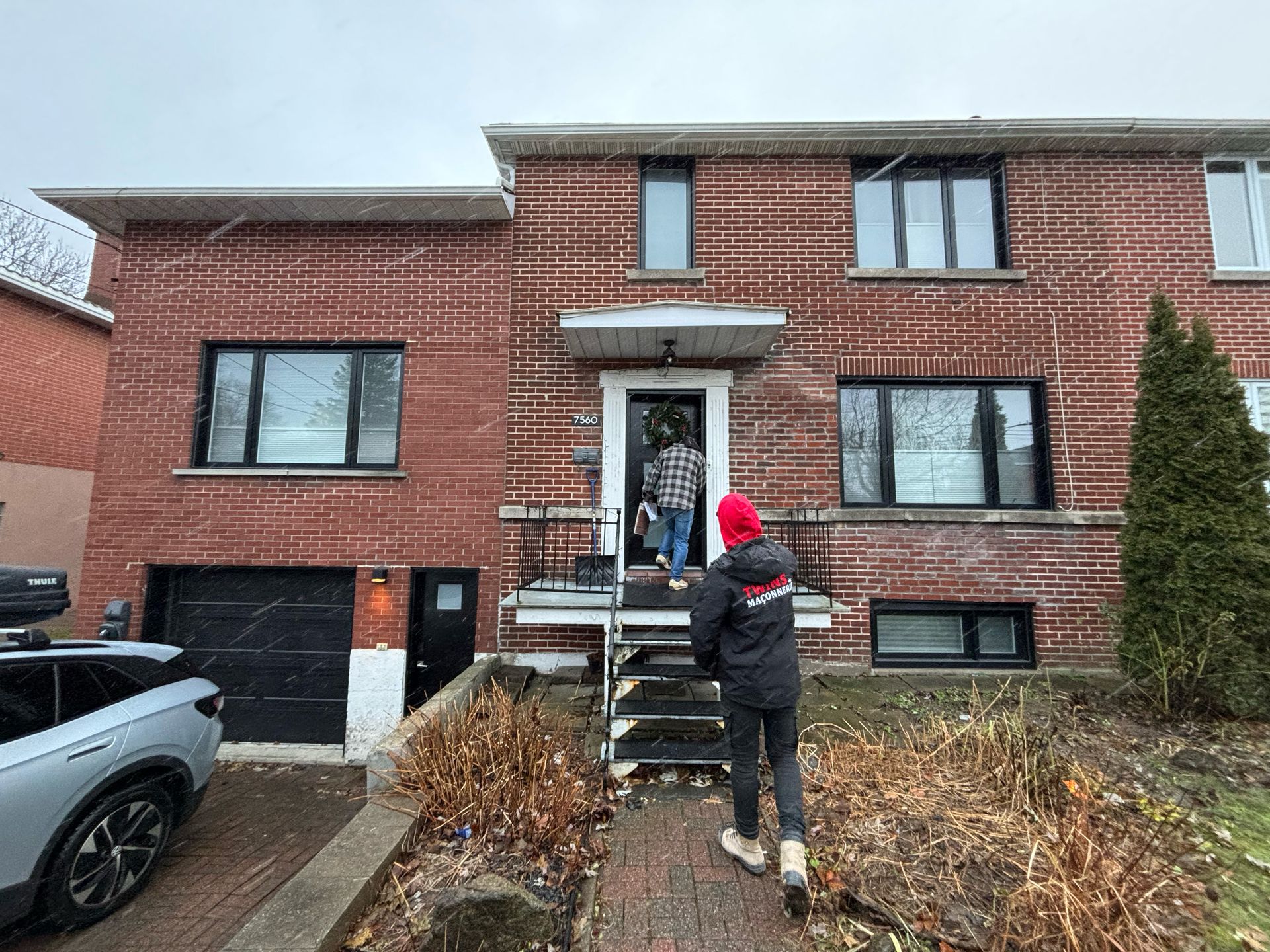 Deux personnes devant une maison en briques rouges, l'une entre, l'autre fait face à la maison. Il neige.