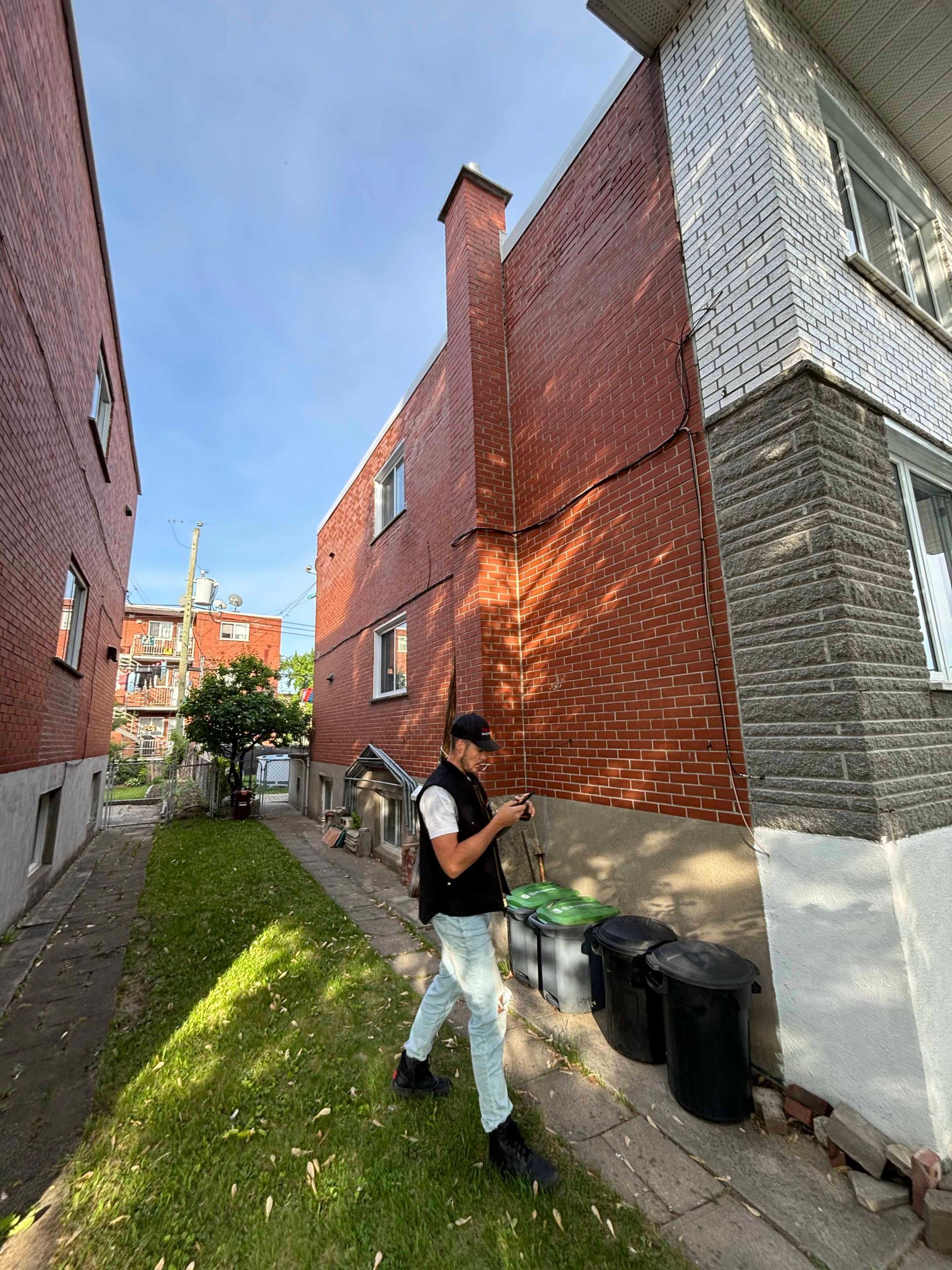 Un homme en jean et gilet se tient près de bâtiments en briques, des poubelles étant posées sur l'herbe sous un ciel bleu.