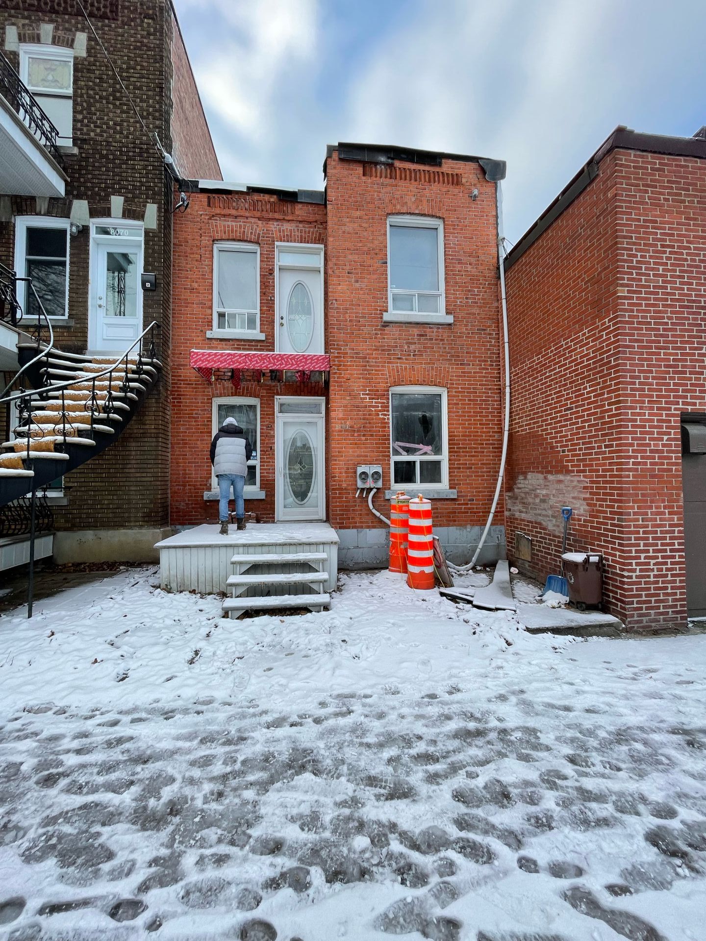 Bâtiment en briques de deux étages sous la neige, une personne sur le porche. Porte blanche, auvent rouge, cônes de signalisation orange.