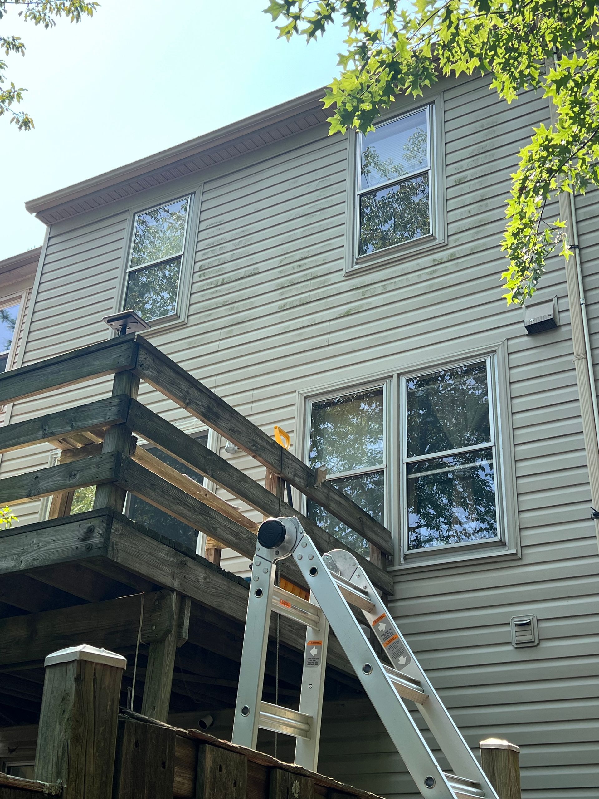 Back of a house with a wooden deck and a ladder leaning against the wall, near windows.