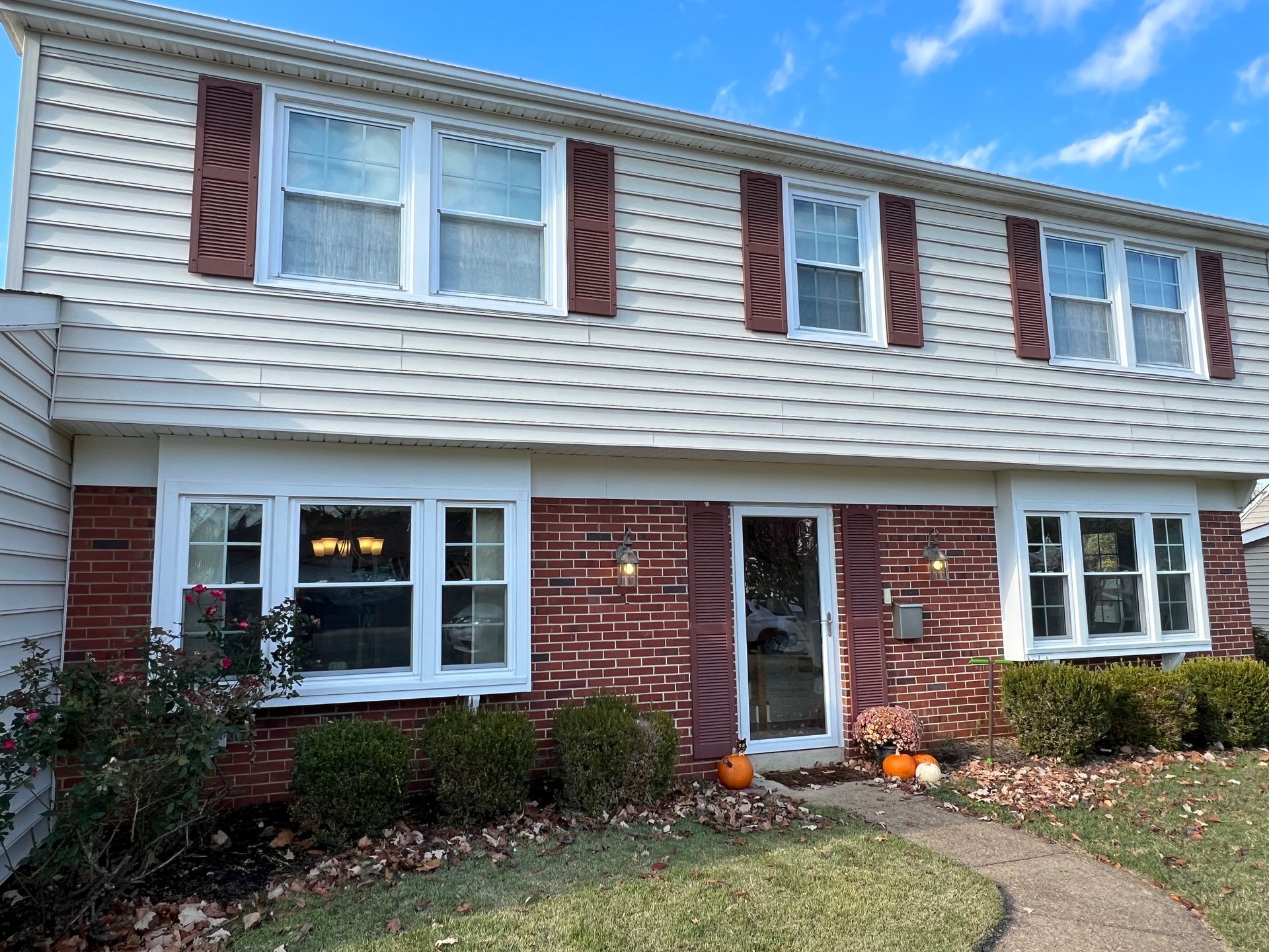 Two-story brick and white house with brown shutters and trim, walkway, and pumpkins.