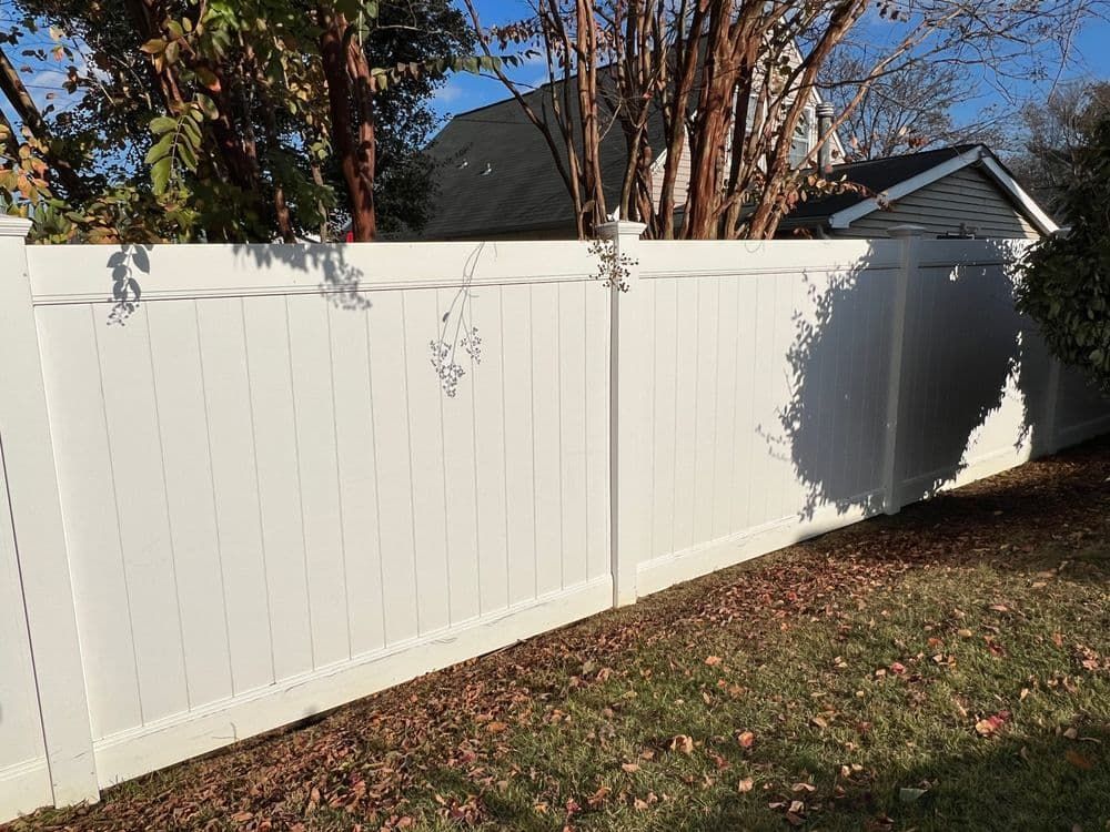 White vinyl fence in a backyard, with foliage and a house visible in the background.