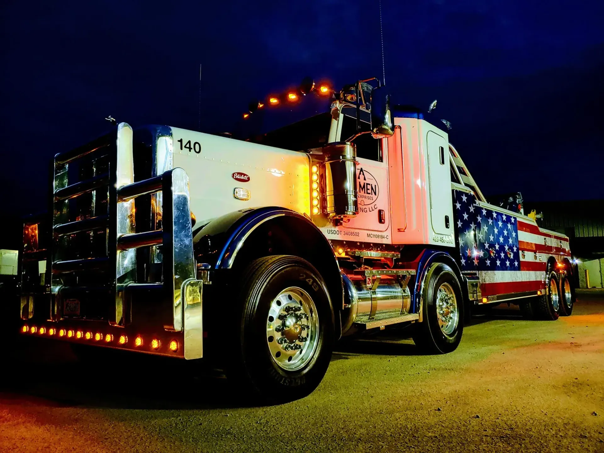 A tow truck with an american flag painted on it is parked in a parking lot at night.