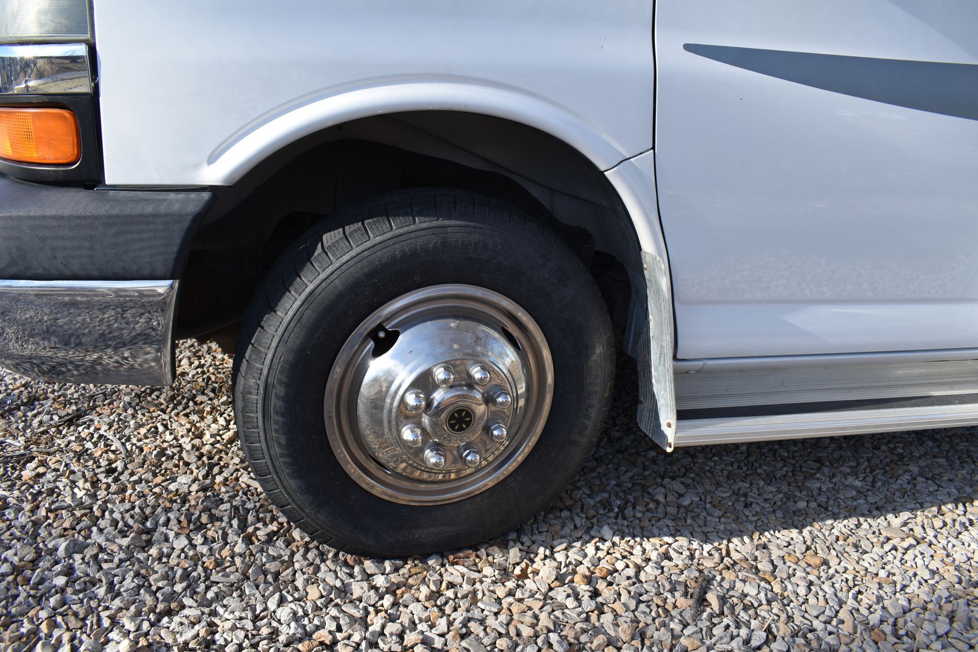 Close-up of a silver van’s front wheel and fender on gravel.