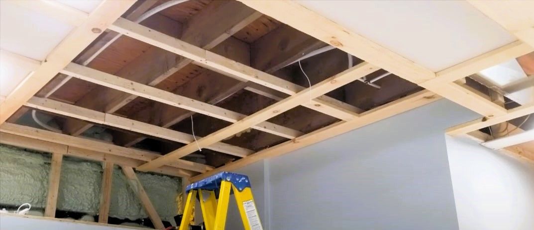 Exposed wooden beams in a building's basement ceiling with partial drywall installation and a yellow stepladder underneath