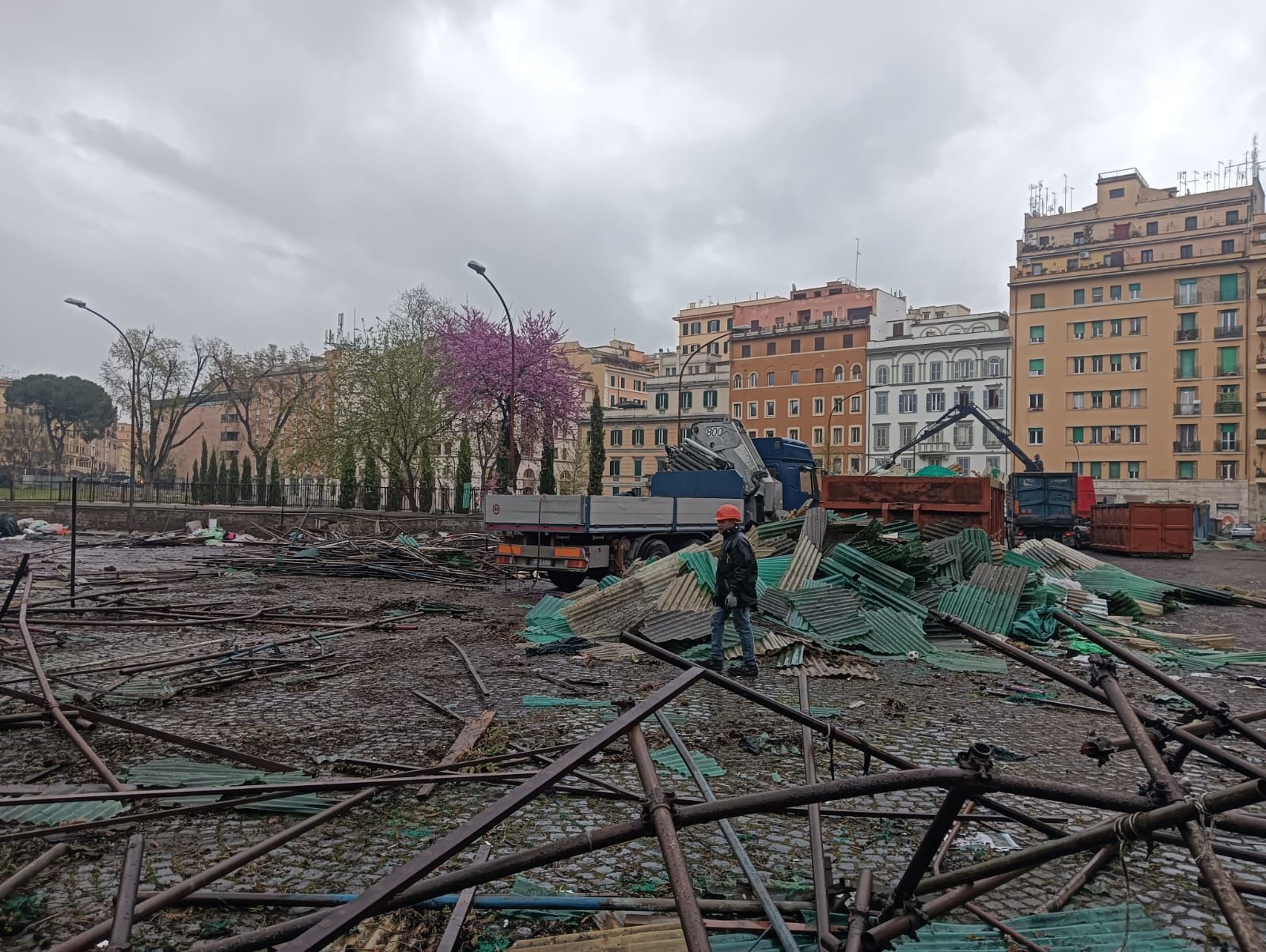 Un escavatore giallo sta demolendo un edificio in cemento, con macerie e polvere in un cantiere.