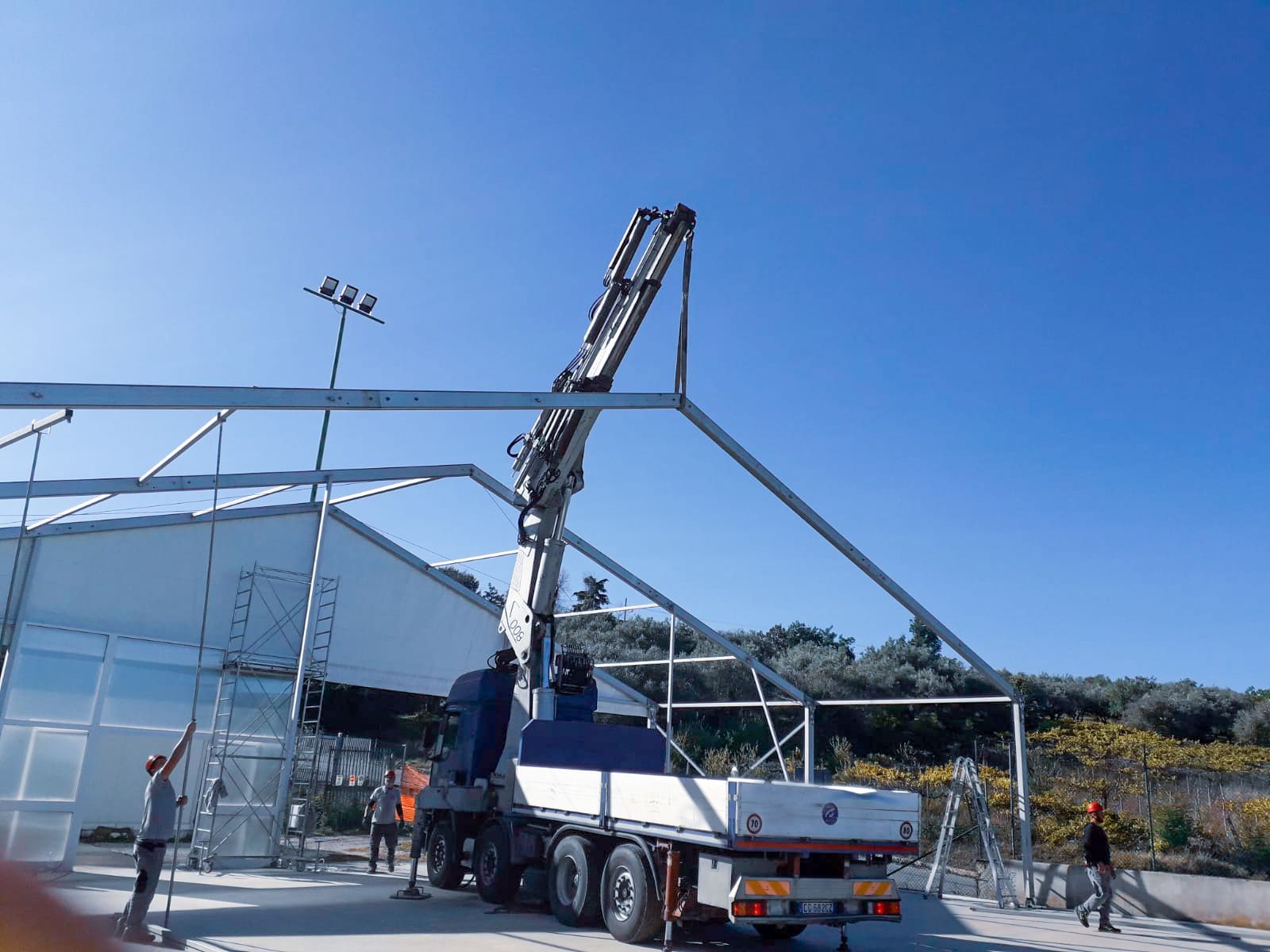 Un camion gru mobile giallo solleva una trave d'acciaio con un gancio in un'area industriale.