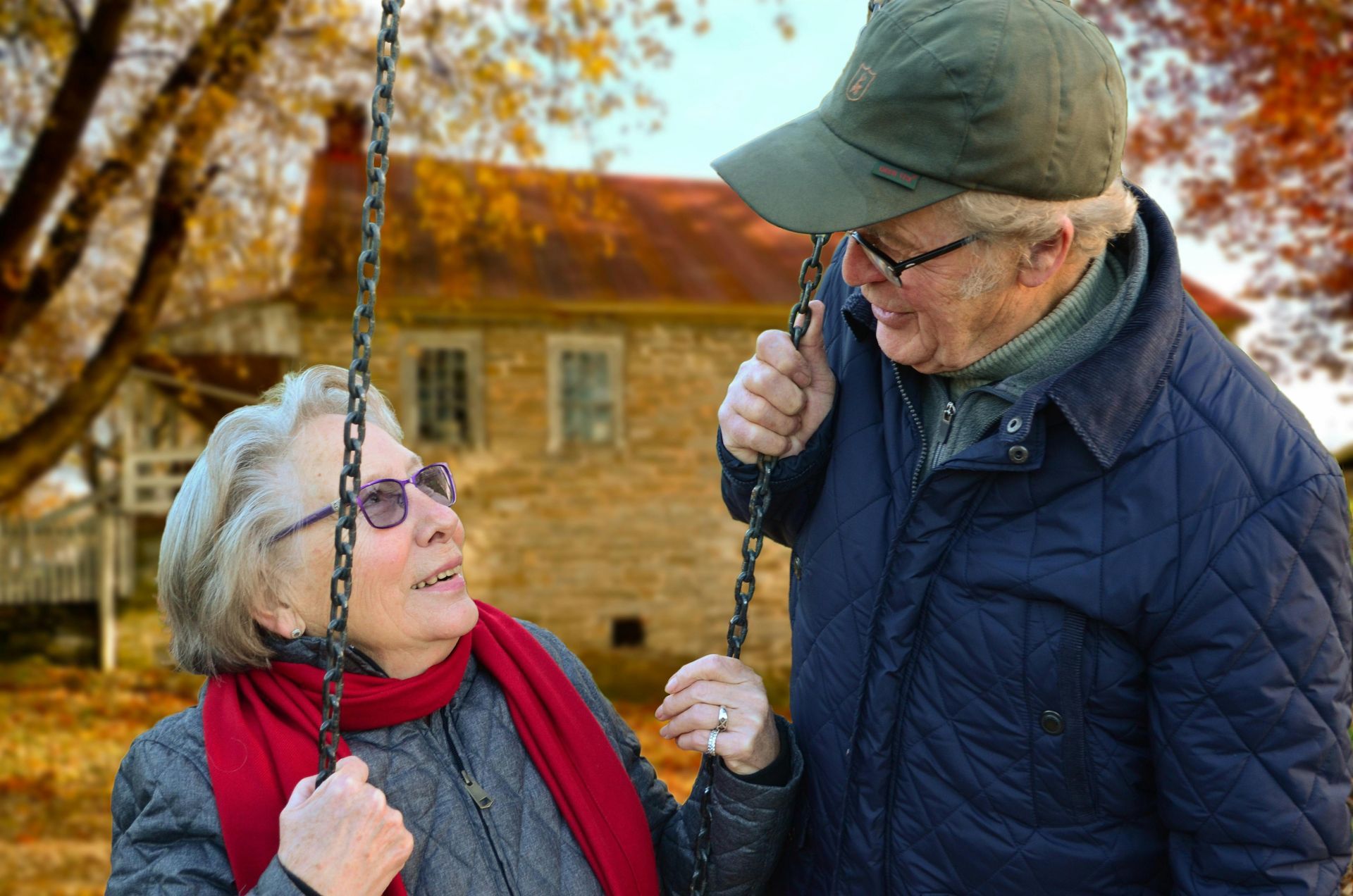 Elderly couple on a swing set, smiling at each other in front of a house in autumn.