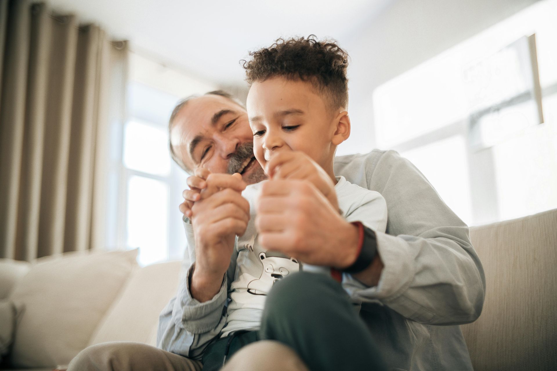 Man and boy laughing together on a couch; the man hugs the boy from behind.
