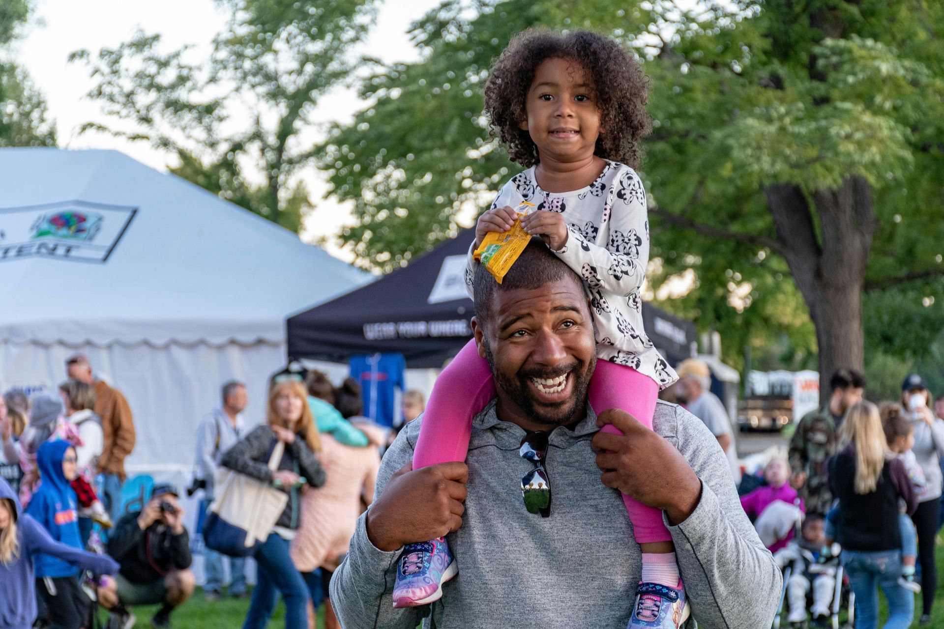 A Black man smiles as he carries a young girl on his shoulders at an outdoor event.