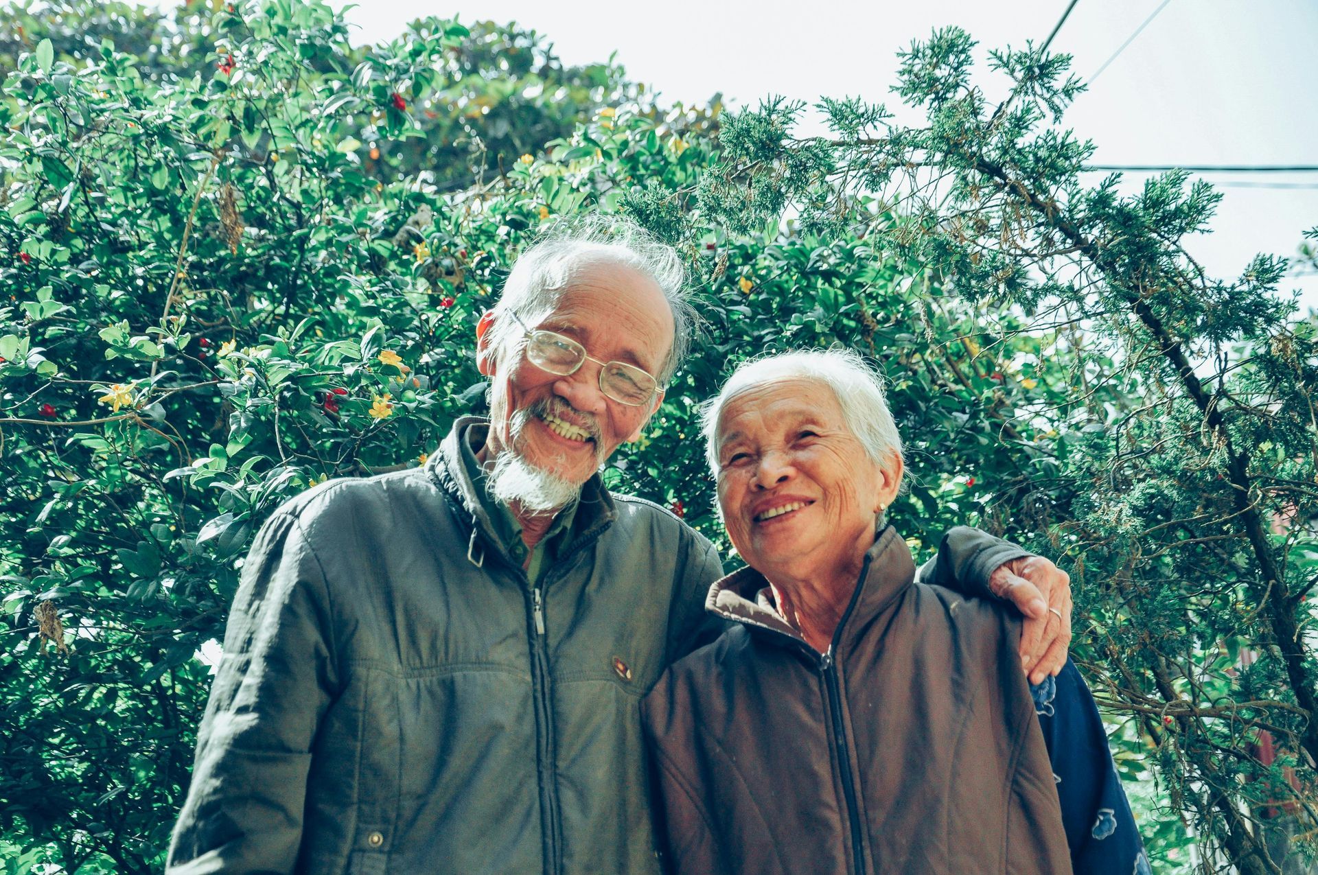 Elderly couple smiling, hugging, posing together outdoors in front of greenery.
