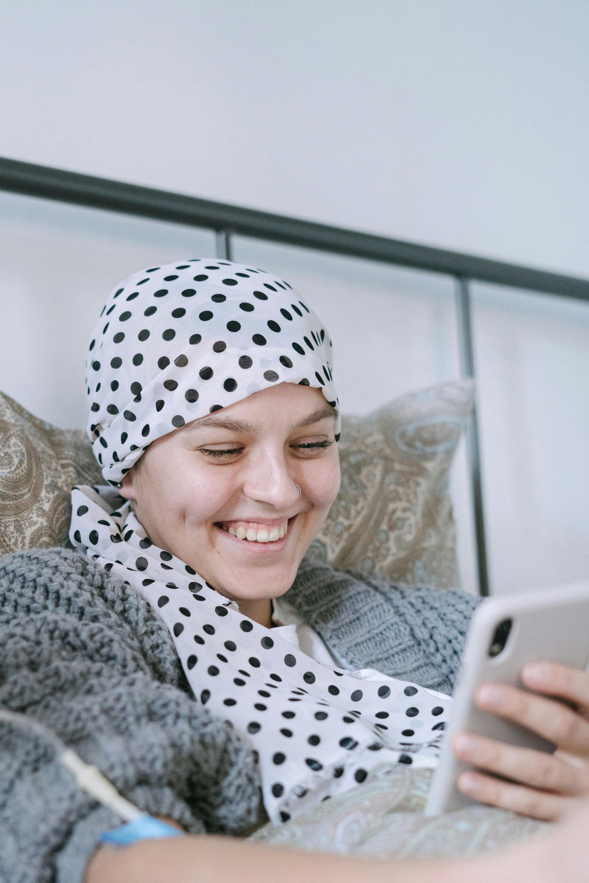 Woman with headscarf smiles while looking at her phone in bed.