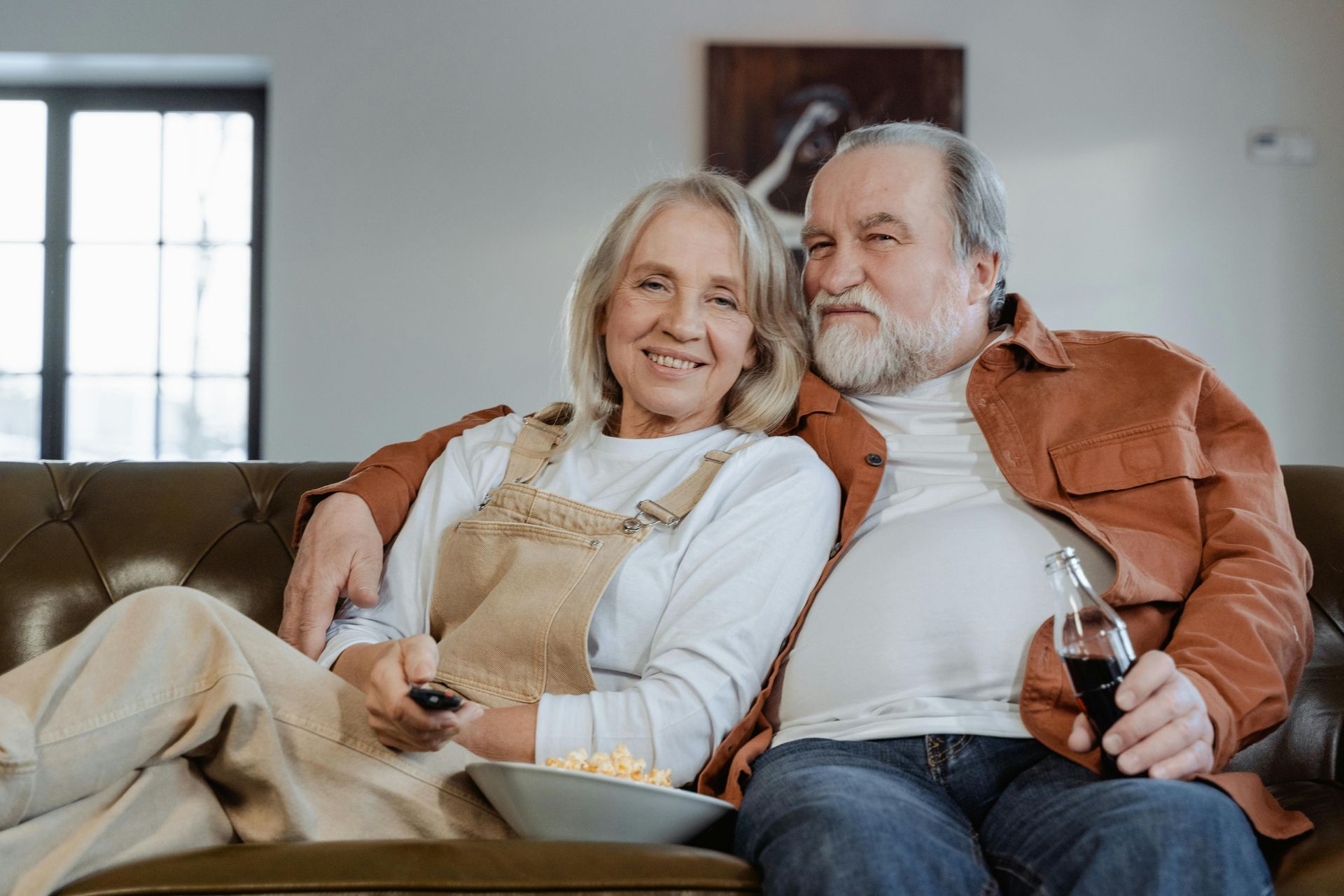 Elderly couple on a couch, watching TV. Woman holds remote, man holds soda. They are smiling.