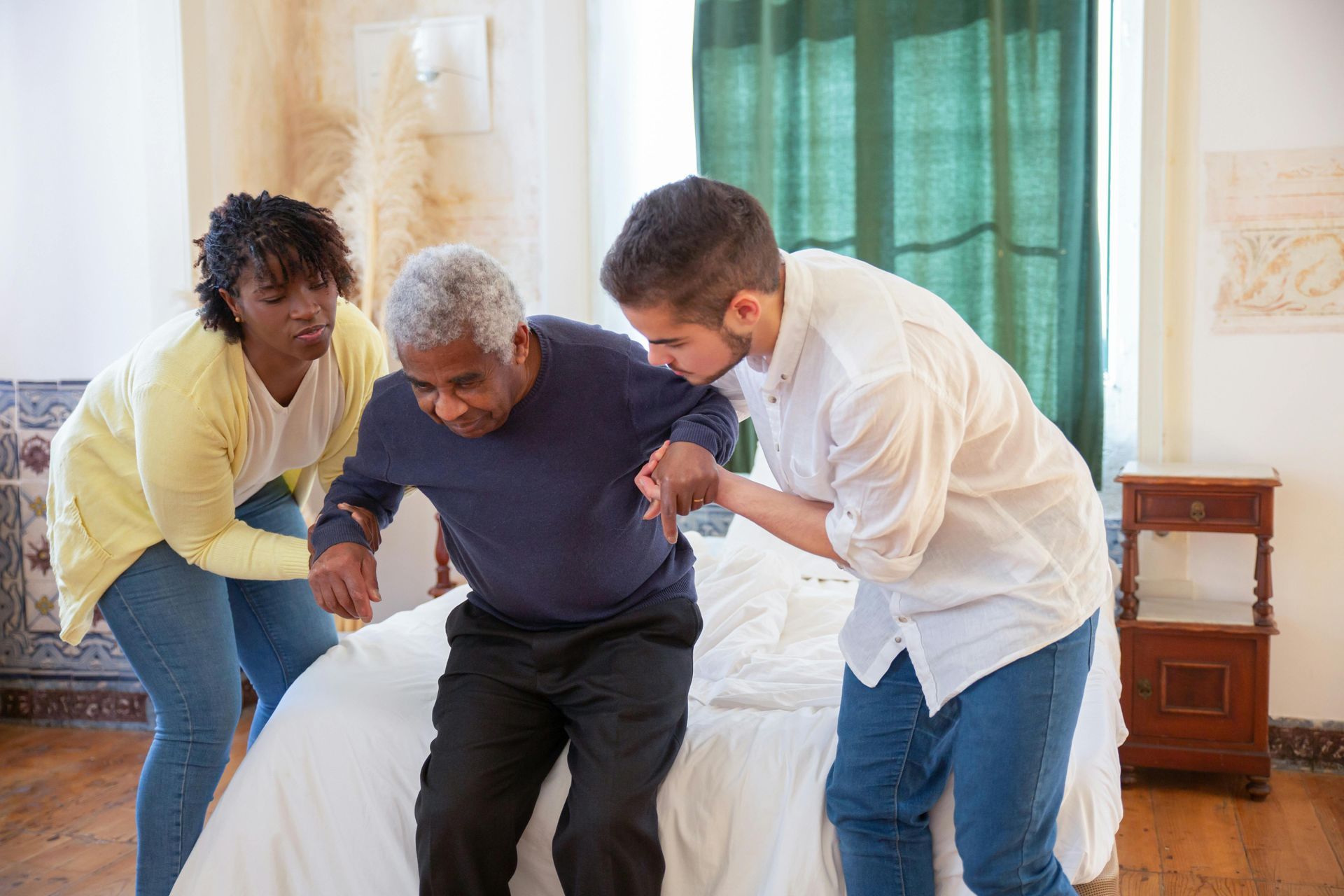Two people assisting an older man to stand up from a bed in a bedroom.