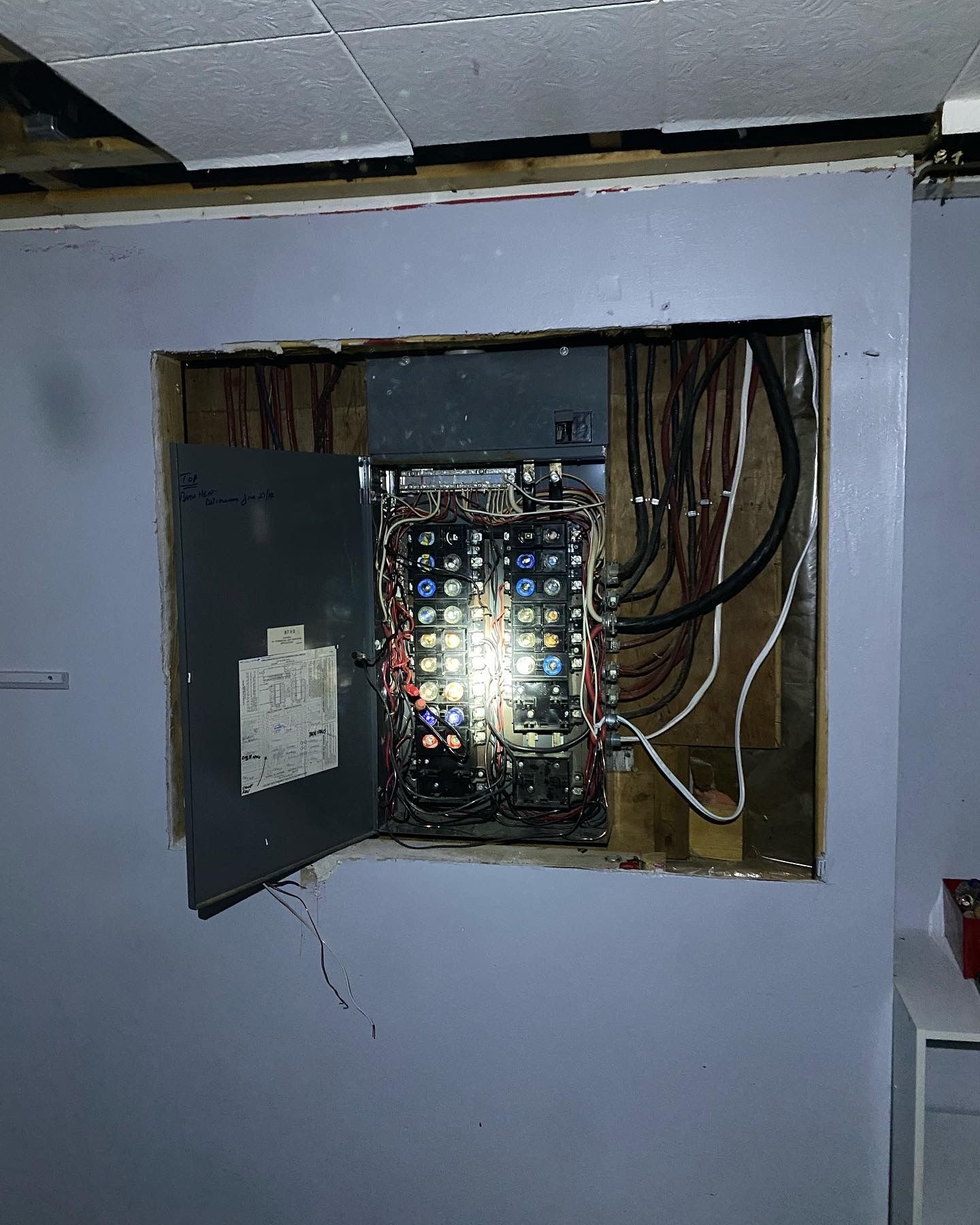 Electrical panel exposed in a wall, wires visible, door open. Gray panel, wood frame, blue wall.