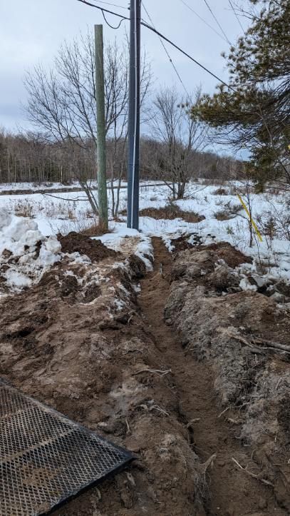Trench in dirt leads toward utility poles, snow on either side, in a wintery outdoor setting.