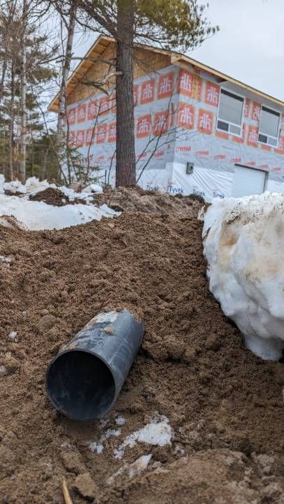 Black pipe emerging from a trench in dirt, in front of a house under construction; snow present.