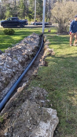 A trench with black pipe next to a grassy lawn. A person stands nearby, a black truck in the background.
