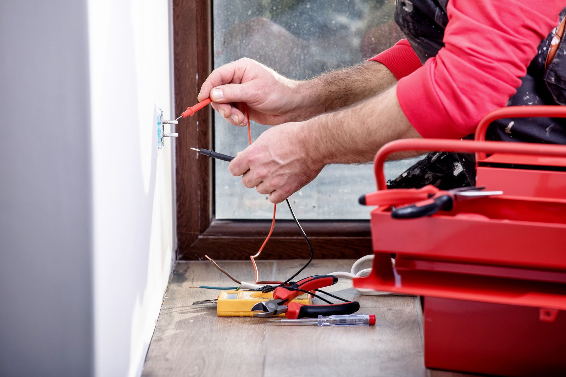 Electrician testing an electrical outlet with a multimeter, tools in a red toolbox nearby.