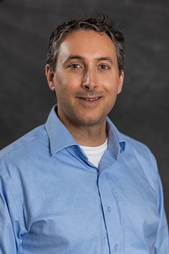 Portrait of a smiling man in a light blue button-down shirt against a gray background